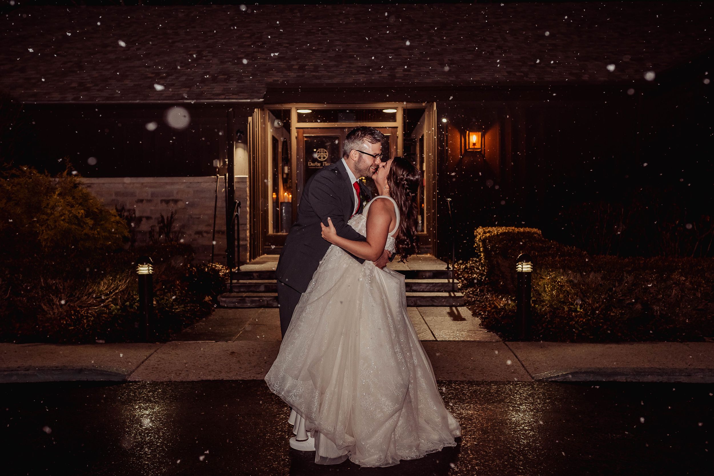 Bride and groom share a romantic kiss in the snow outside a warmly lit venue at night.