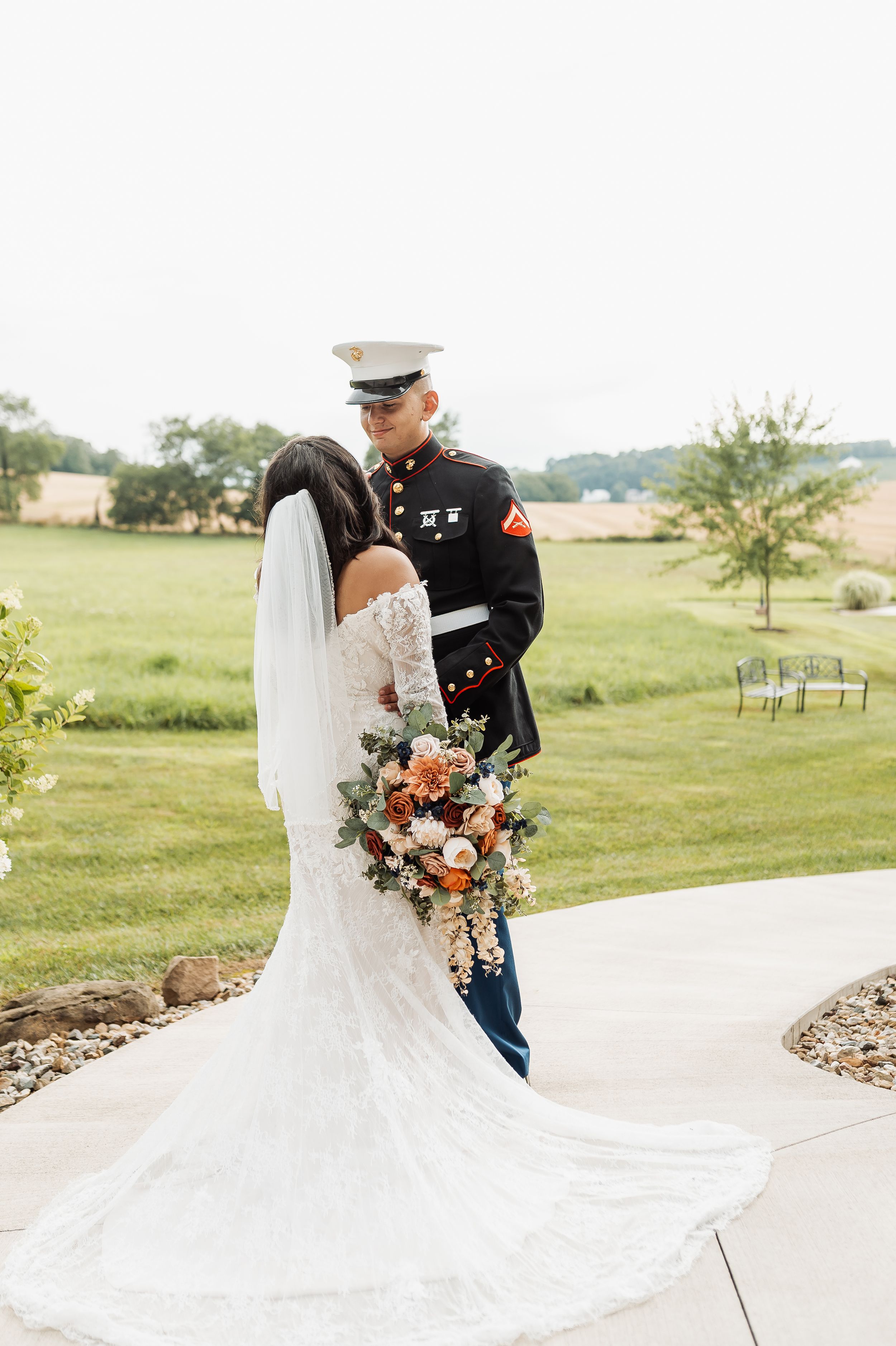 Bride in lace gown with floral bouquet shares first look with Marine groom in dress uniform outdoors.