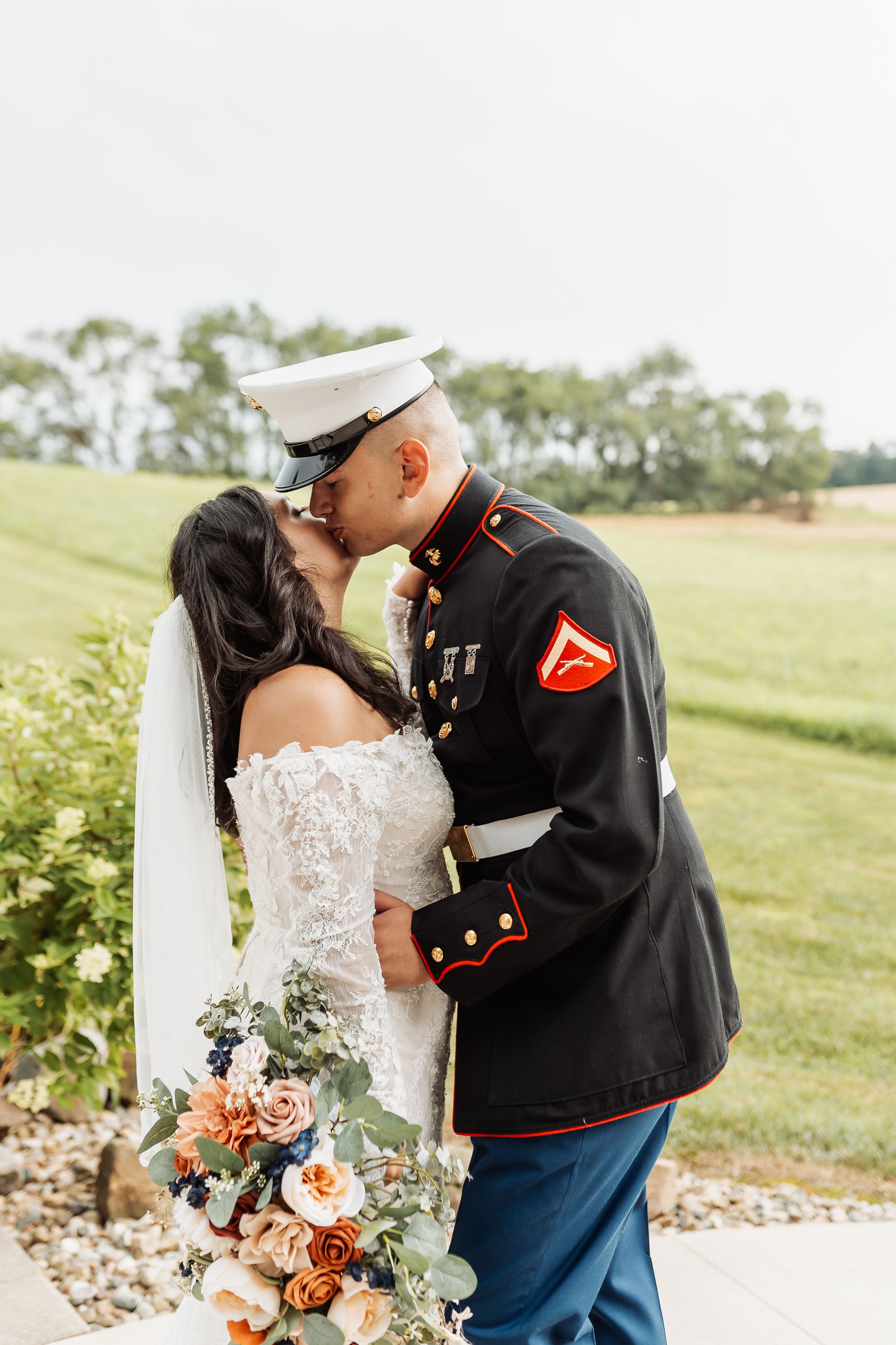 A bride and Marine groom share a kiss outdoors, she holds a floral bouquet in a lace gown as he wears dress blues.