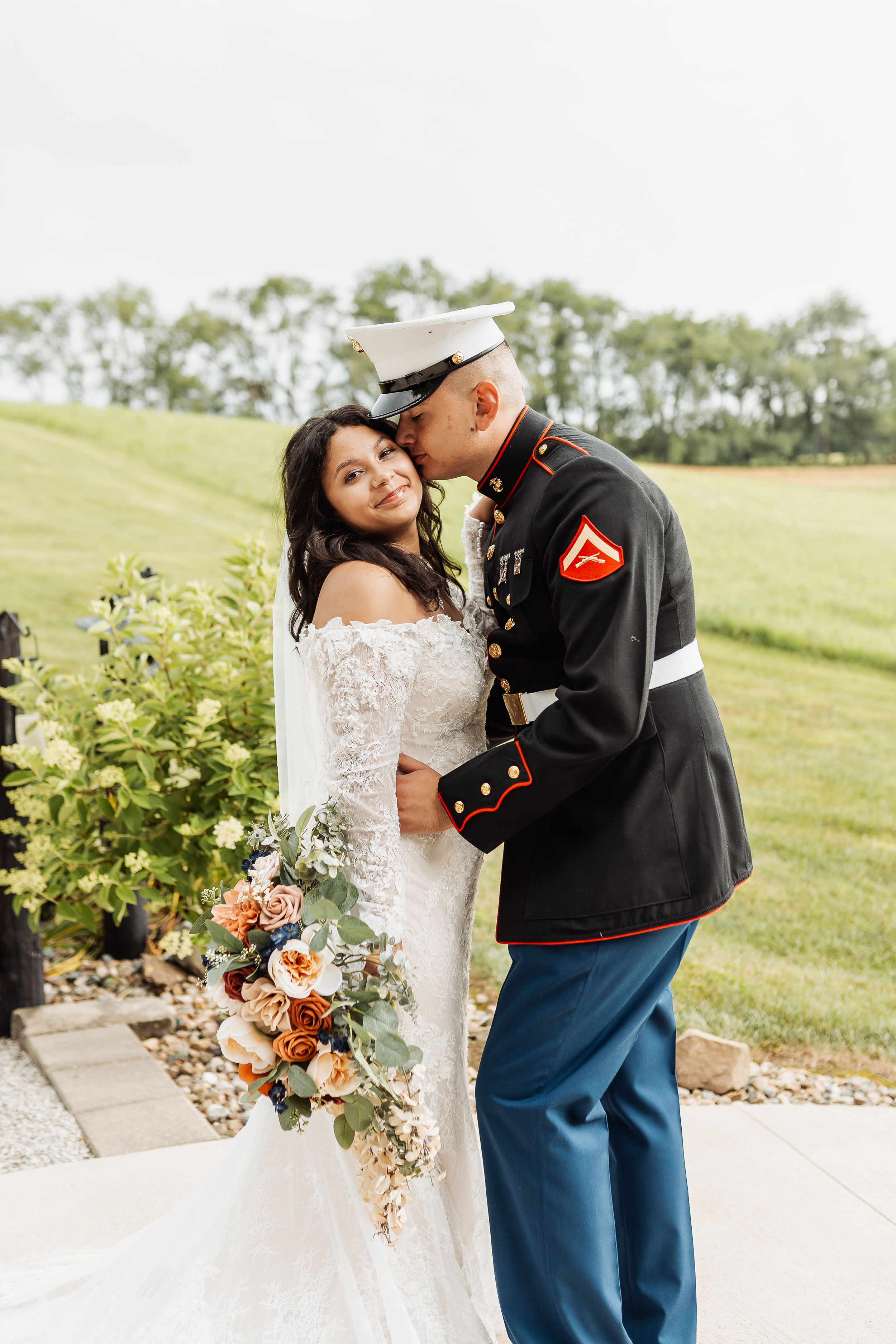 A Marine in dress uniform kisses his bride in a lace gown holding a floral bouquet outdoors.