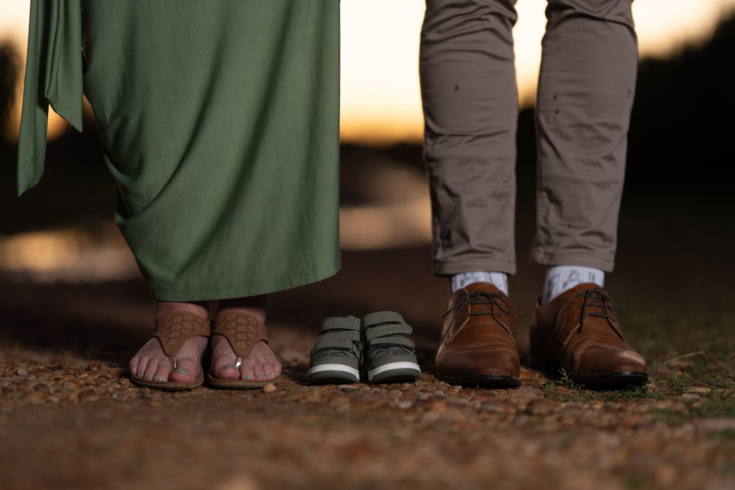 Close-up of three pairs of feet showing parents and tiny baby shoes at golden sunset.