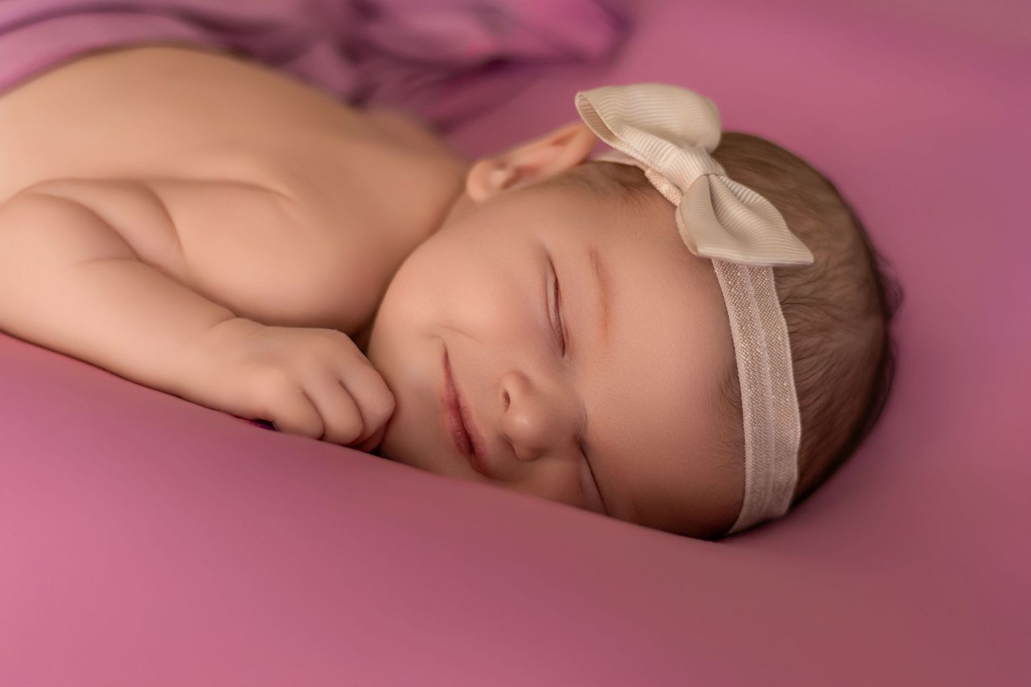 Sleeping newborn with cream bow headband on pink backdrop.