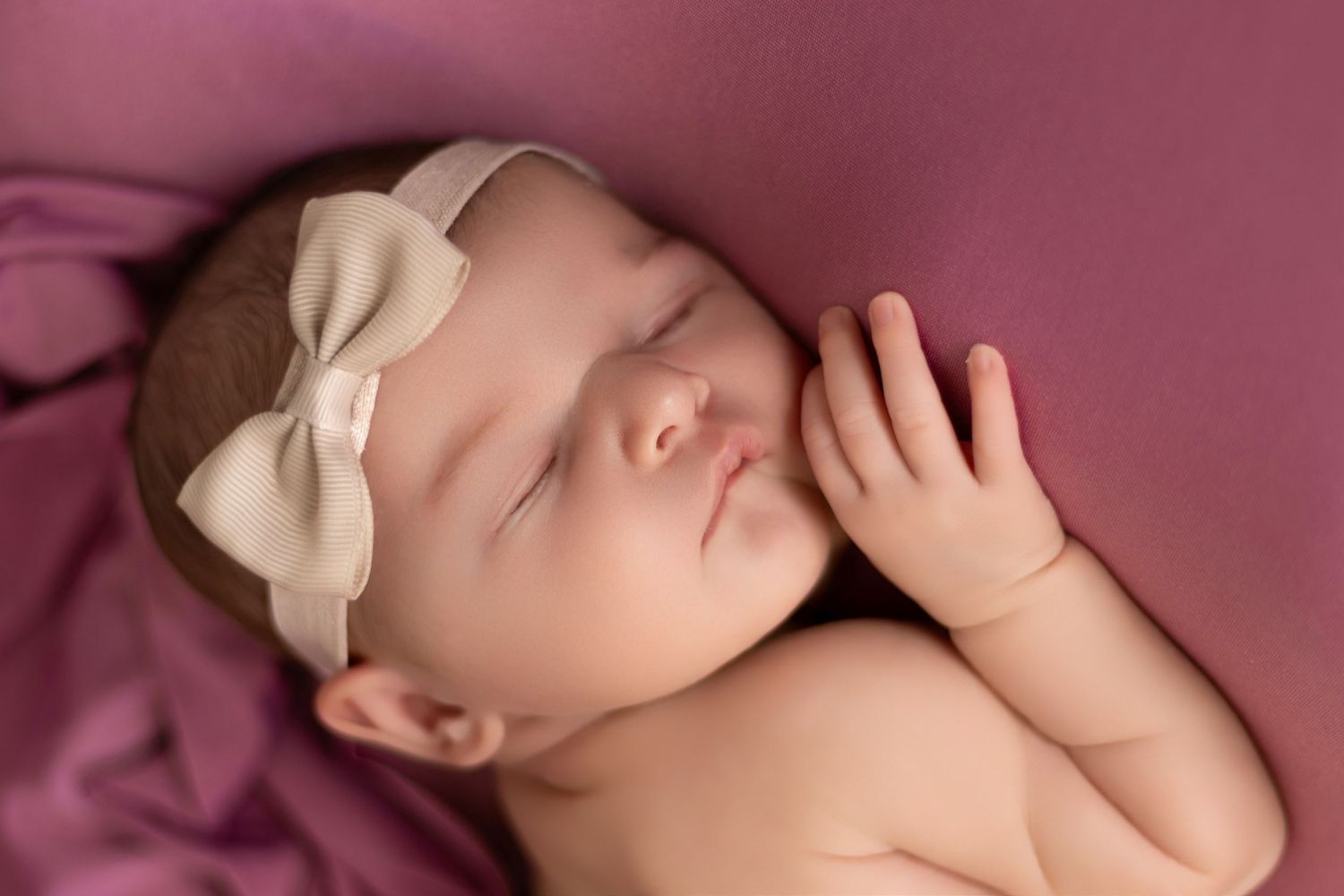 Peaceful newborn resting on soft pink fabric with delicate bow accessory.