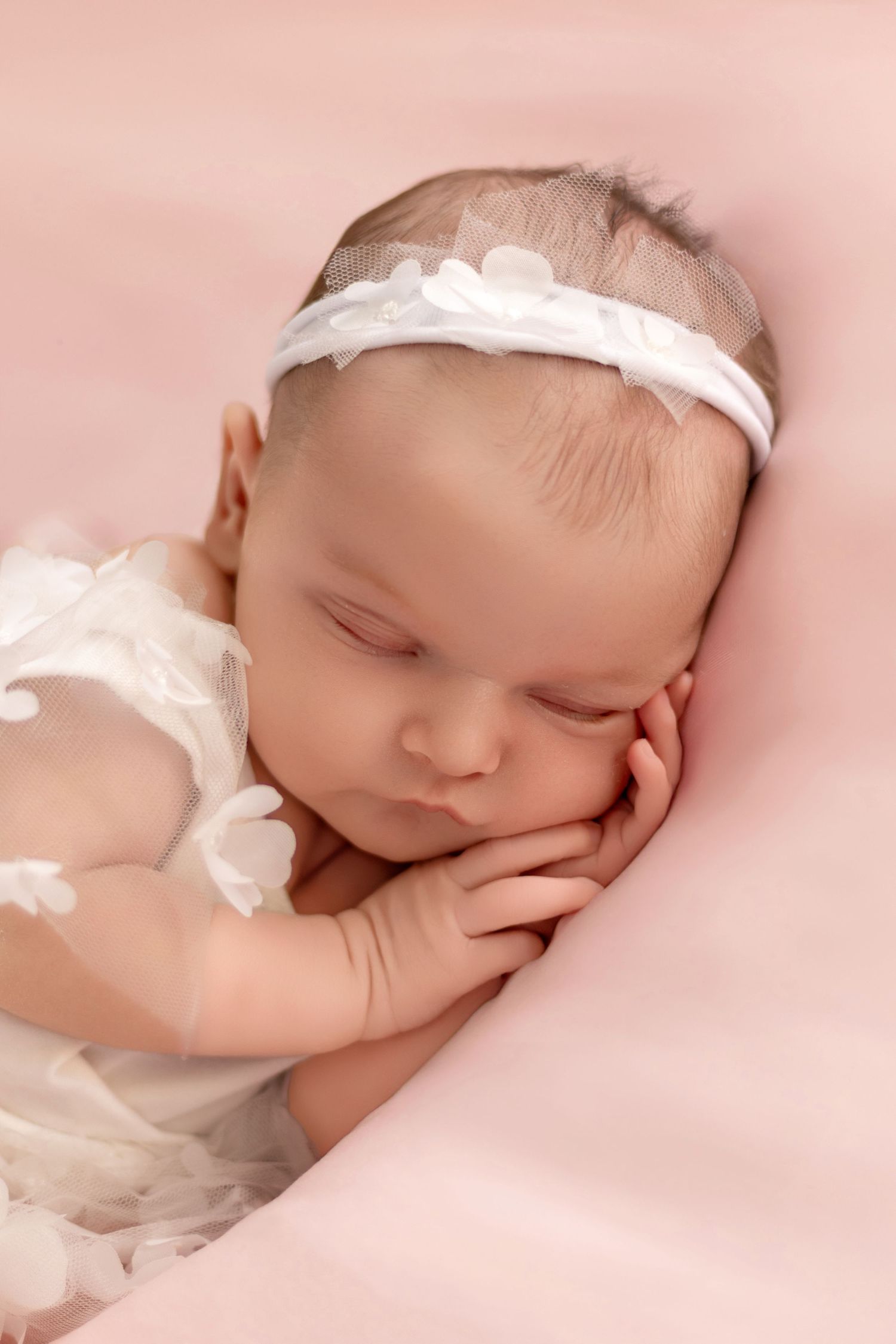 Sleeping newborn with white headband on pink background in artistic pose.