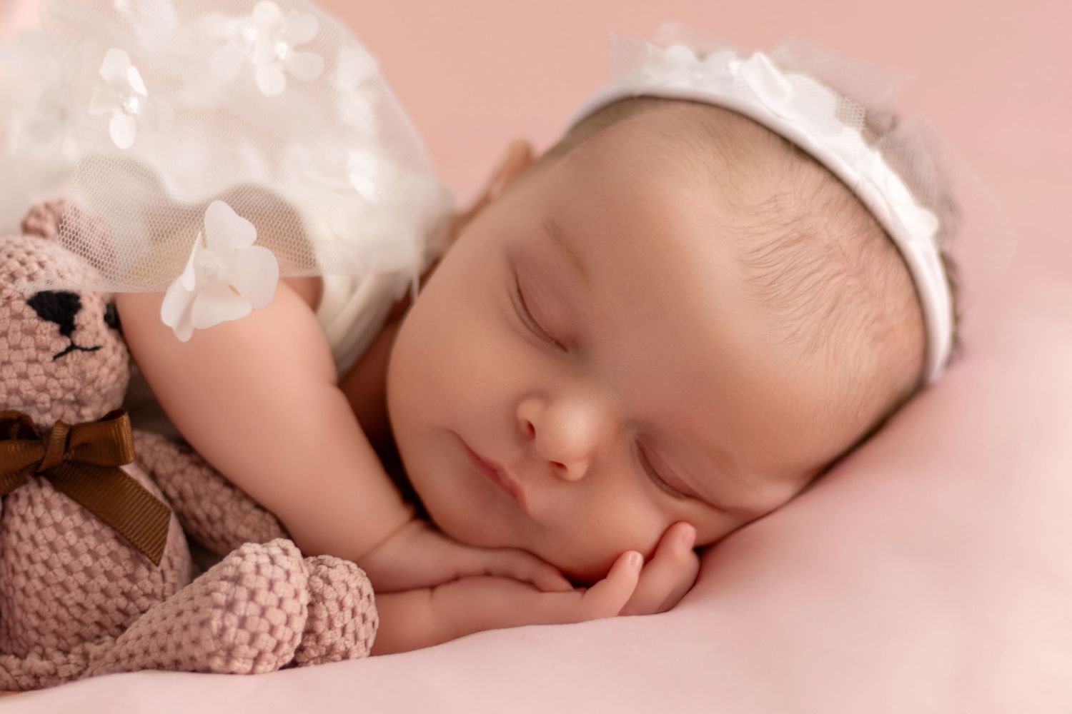Peaceful sleeping newborn with white bow accessory on pink backdrop.