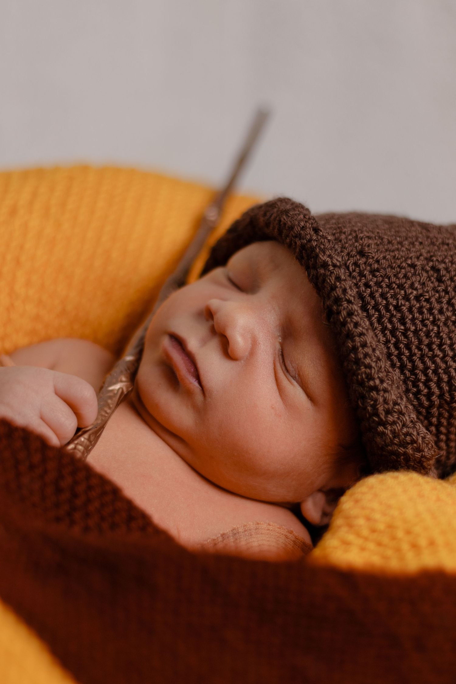 Peaceful newborn sleeping on yellow blanket wearing brown knit hat in warm lighting.