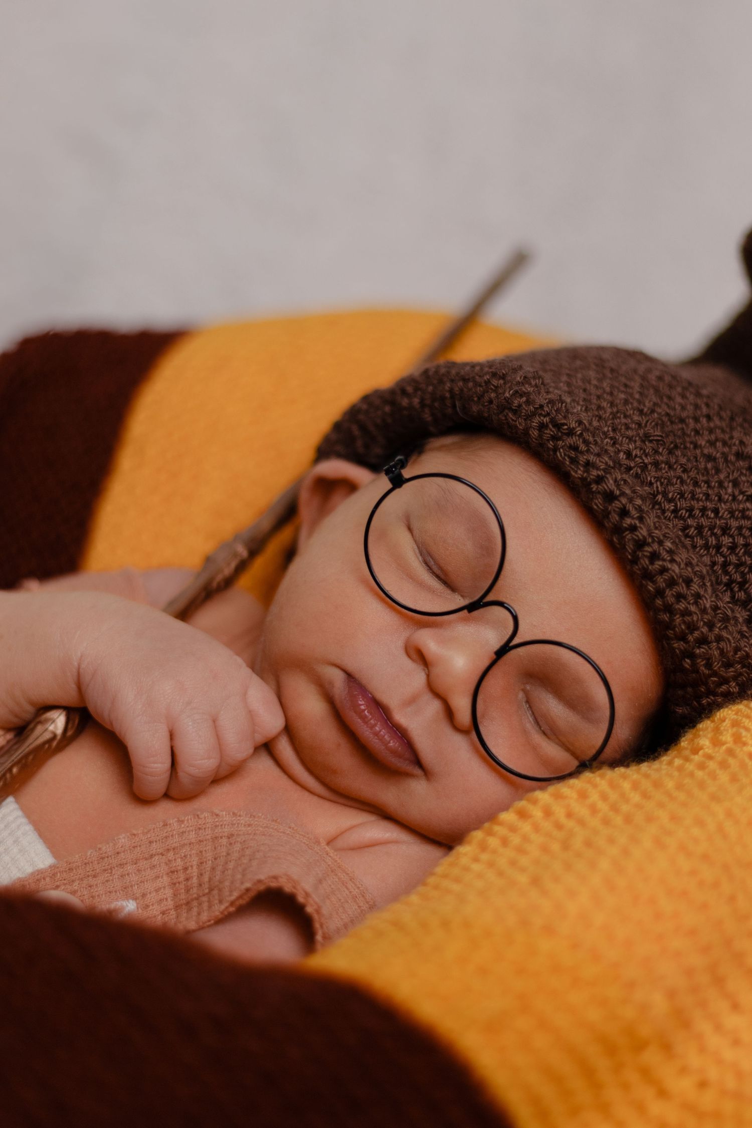 Newborn sleeping peacefully with round glasses and brown knit hat on yellow blanket.