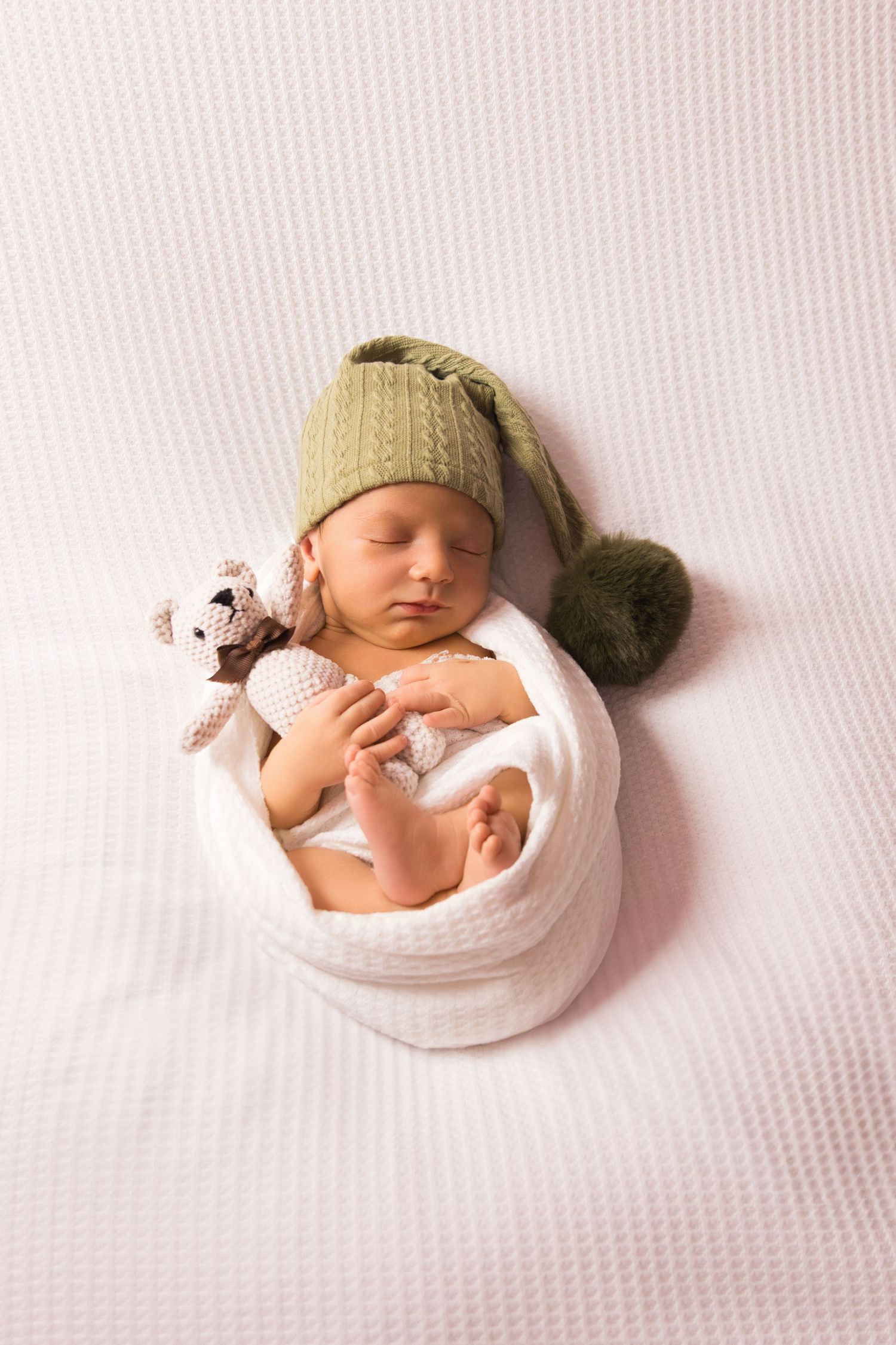 Sleeping newborn wrapped in white blanket wearing green knit hat with small stuffed animal.