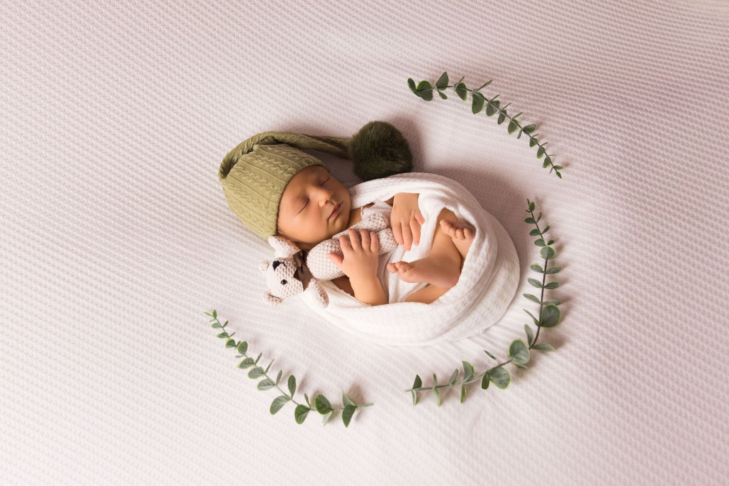 Newborn in white wrap and green hat surrounded by eucalyptus leaves on white backdrop.