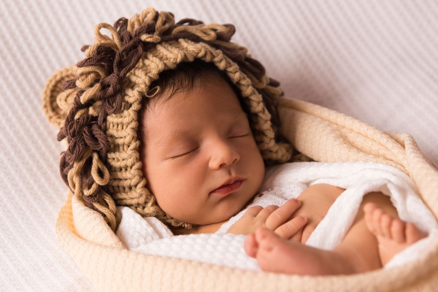Sleeping newborn in knitted lion mane bonnet wrapped in white blanket.