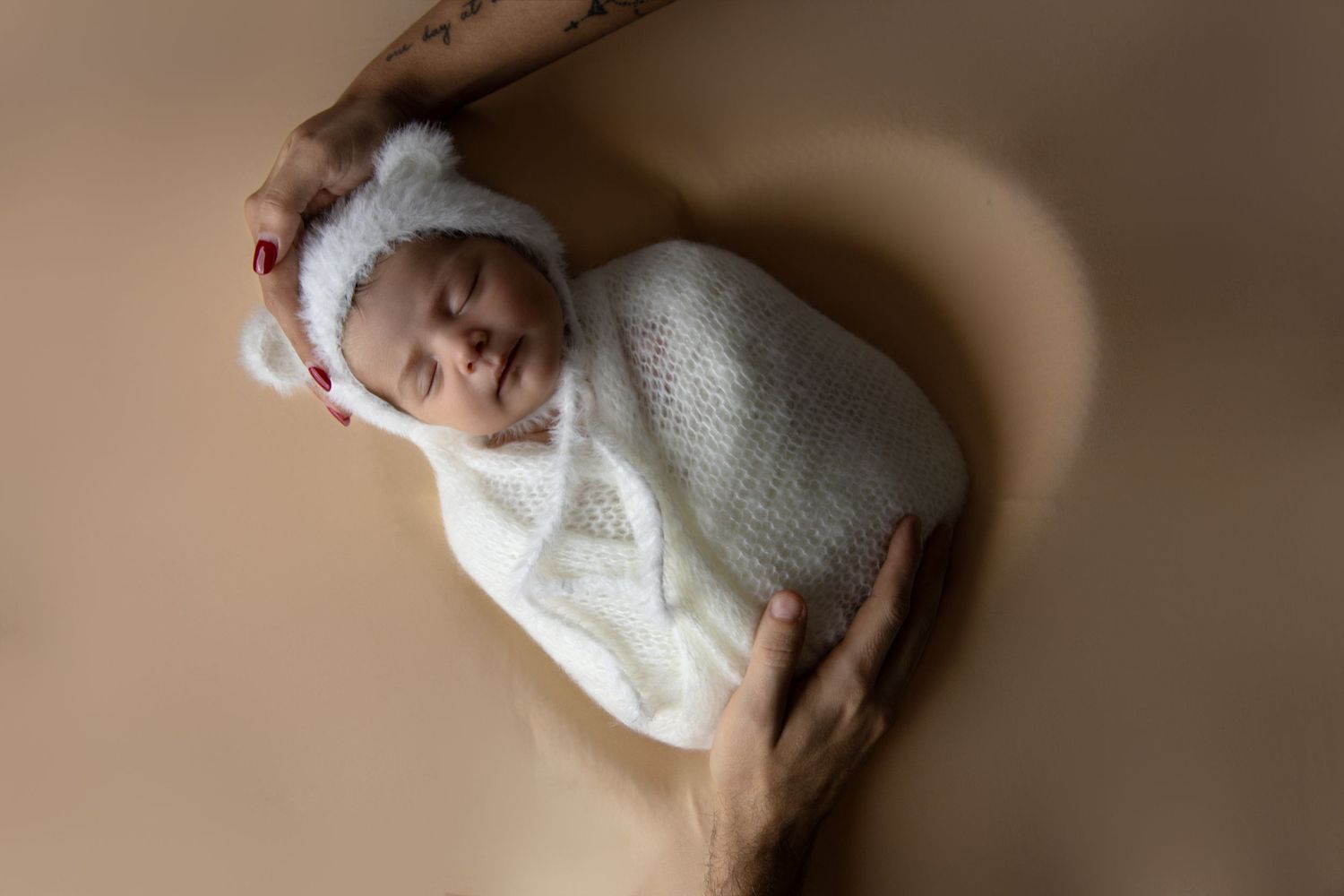 A sleeping baby wearing a white knitted sweater lies peacefully on a beige surface in soft lighting.