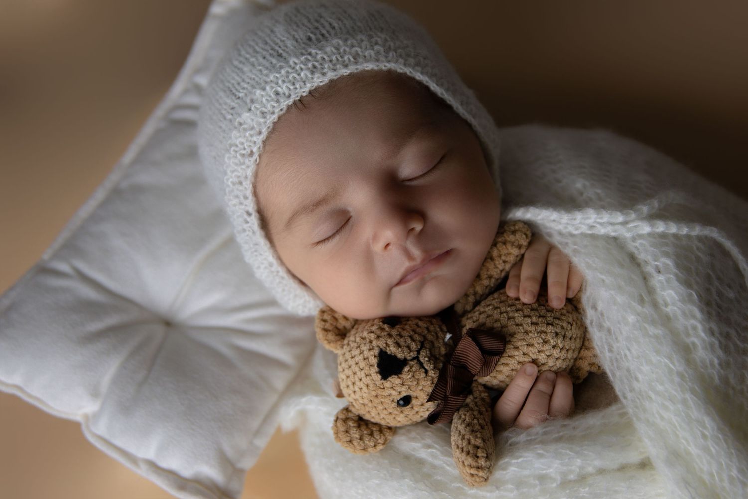 Newborn baby sleeping peacefully on white bedding with a plush teddy bear.