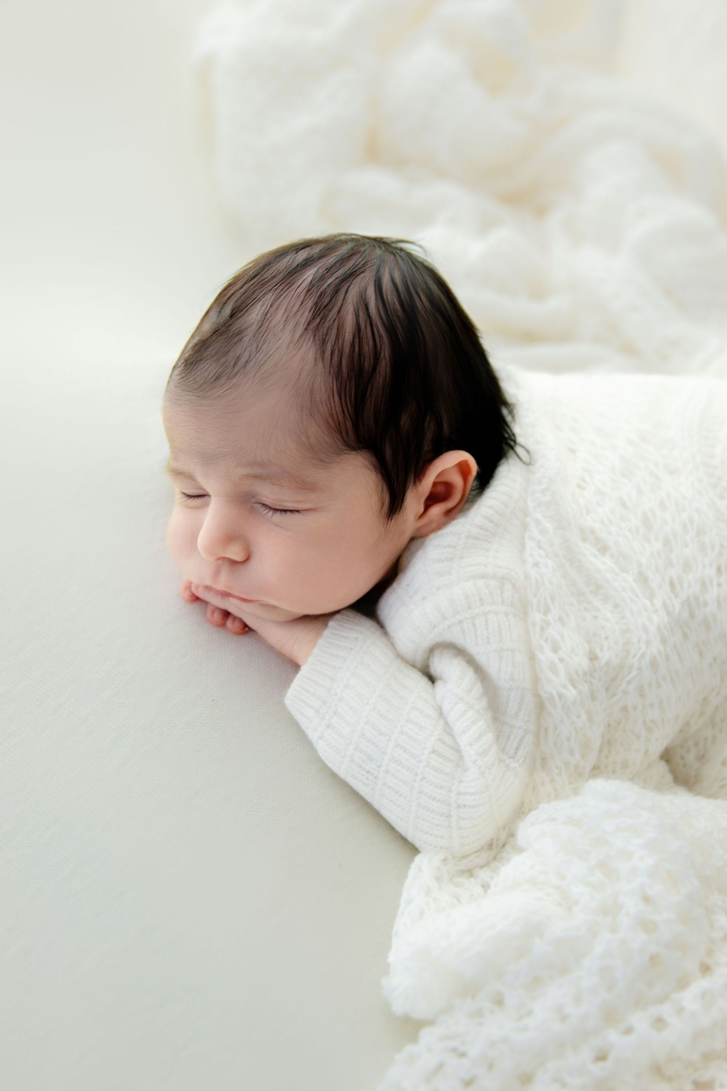 A sleeping newborn baby wrapped in soft white lace blankets rests peacefully against a light background.