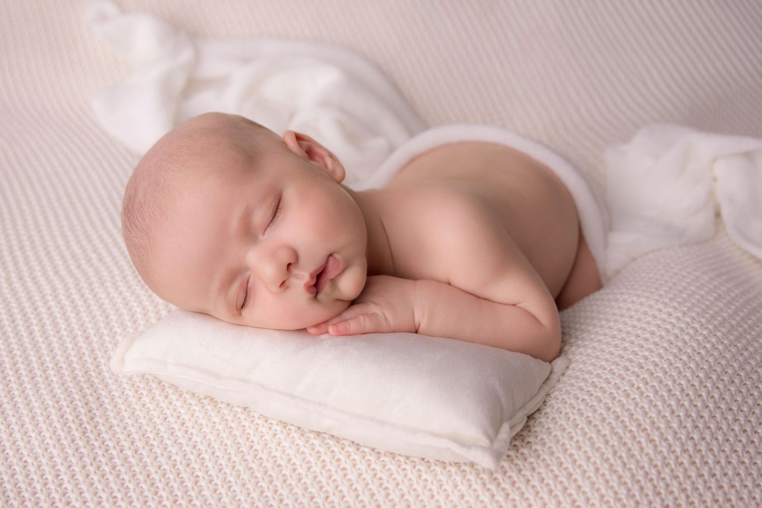 Newborn baby sleeping peacefully on soft white textured blanket in natural light.