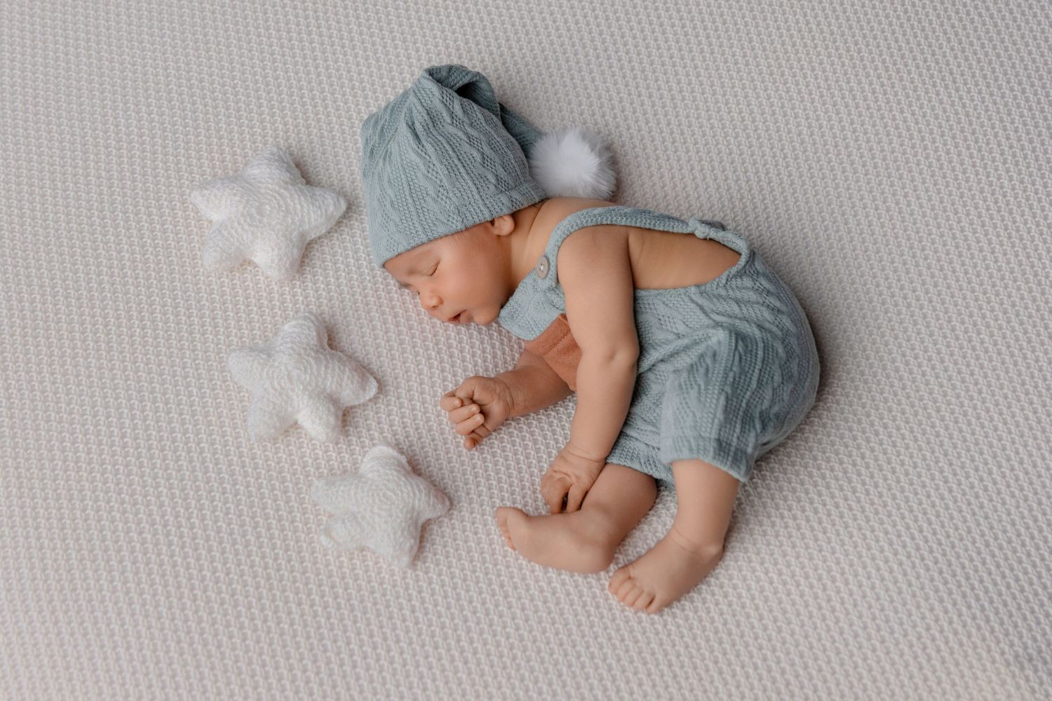 Bebê dorme, vestido com macacão e chapéu azuis, está deitado sobre uma superfície branca texturizada com pequenas estrelas decorativas.