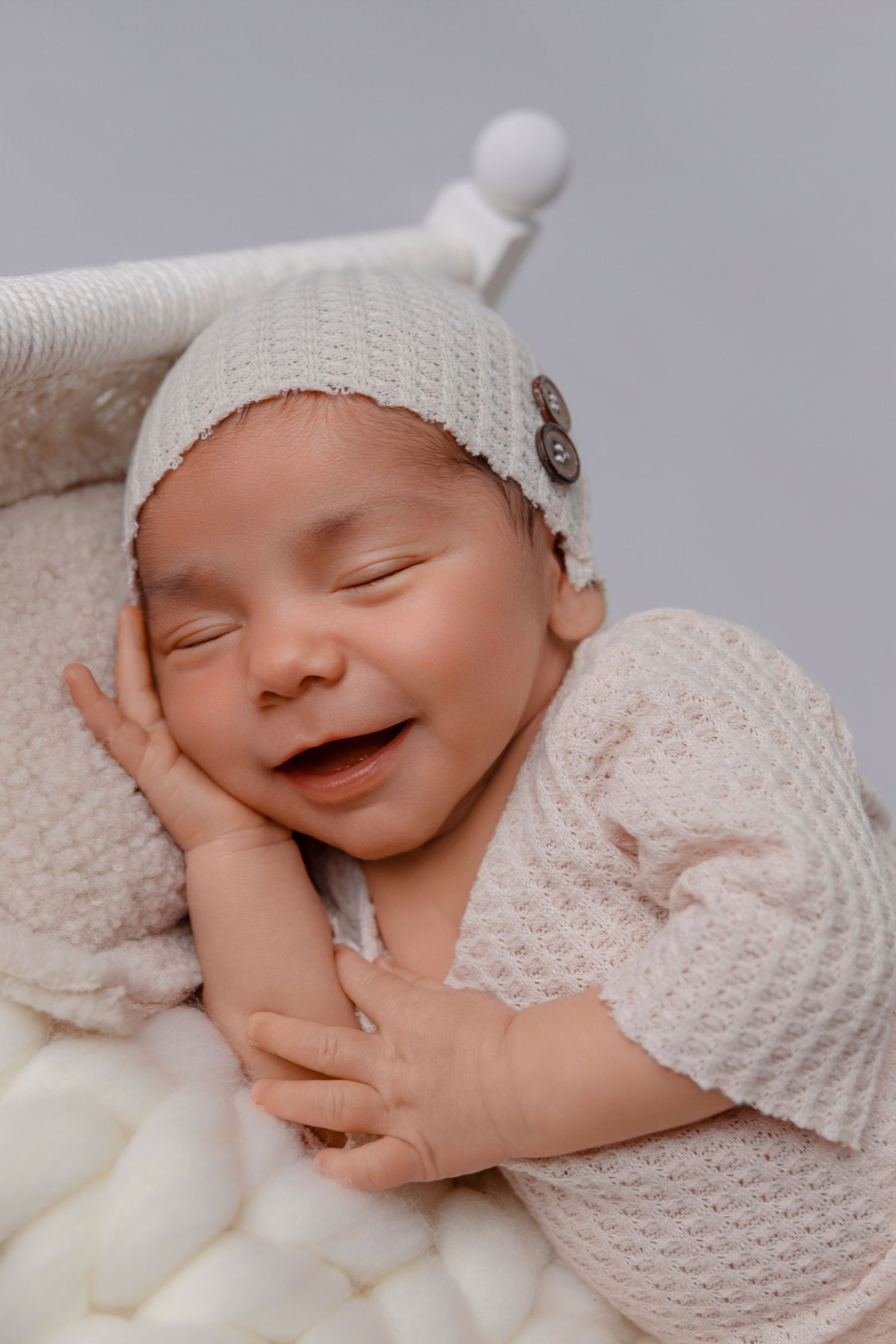 Um bebê recém-nascido, usan gorro e pijama brancos, sorri enquanto dorme numa caminha de bebé de cor creme.