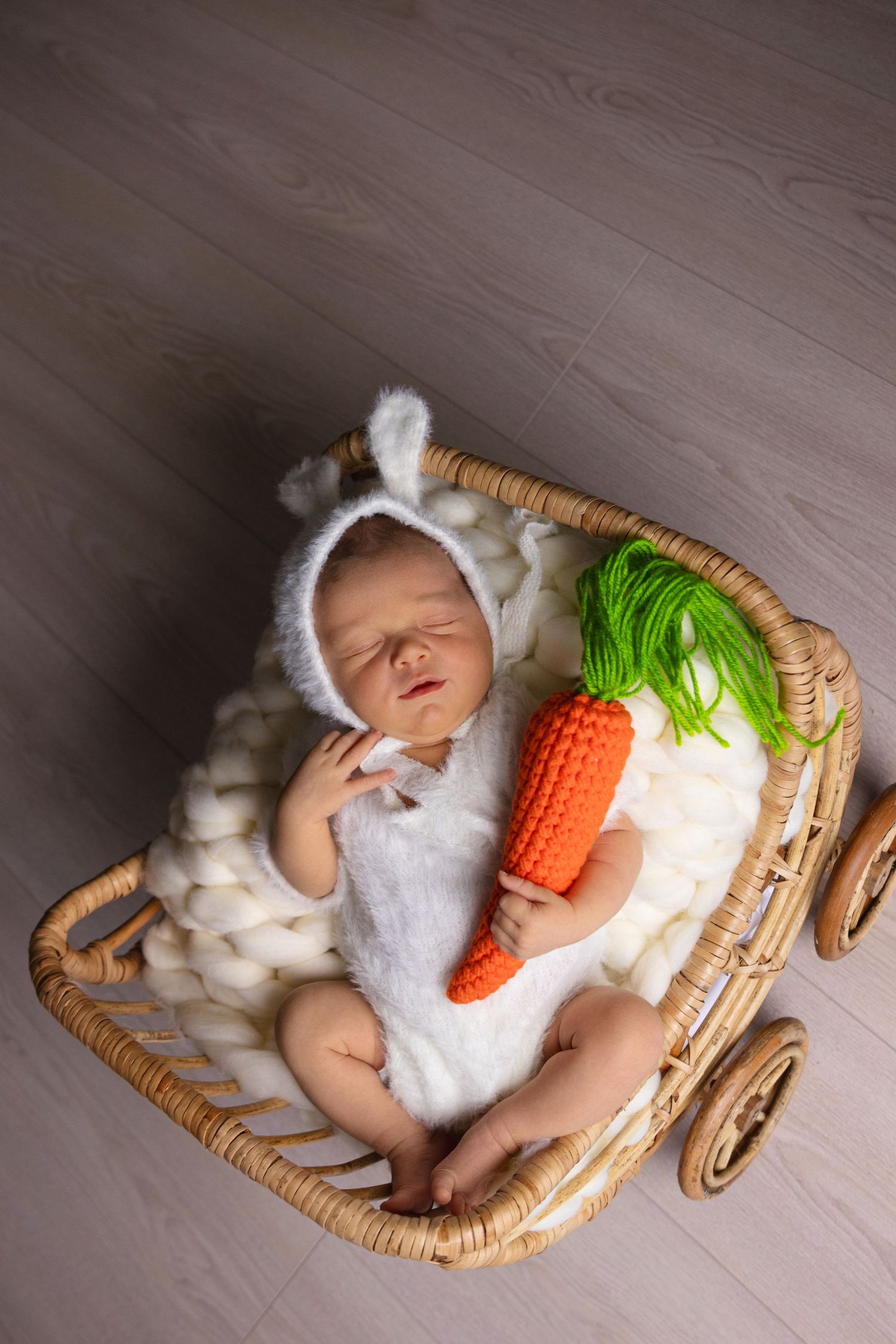 Newborn baby sleeping in wicker carriage with white bunny outfit and decorative carrot prop.