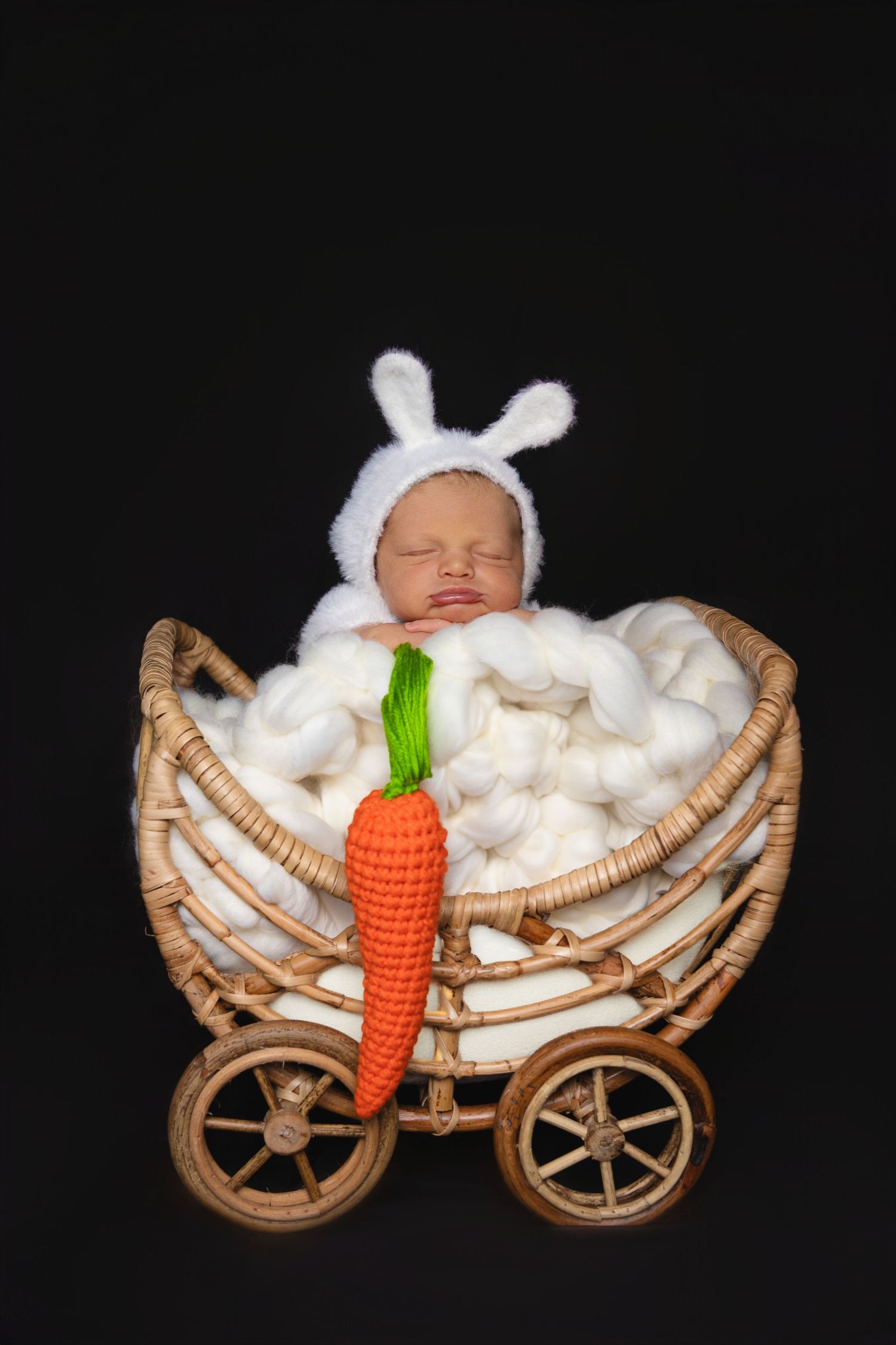 Baby in bunny costume sleeping in wicker carriage with orange carrot prop against dark background.