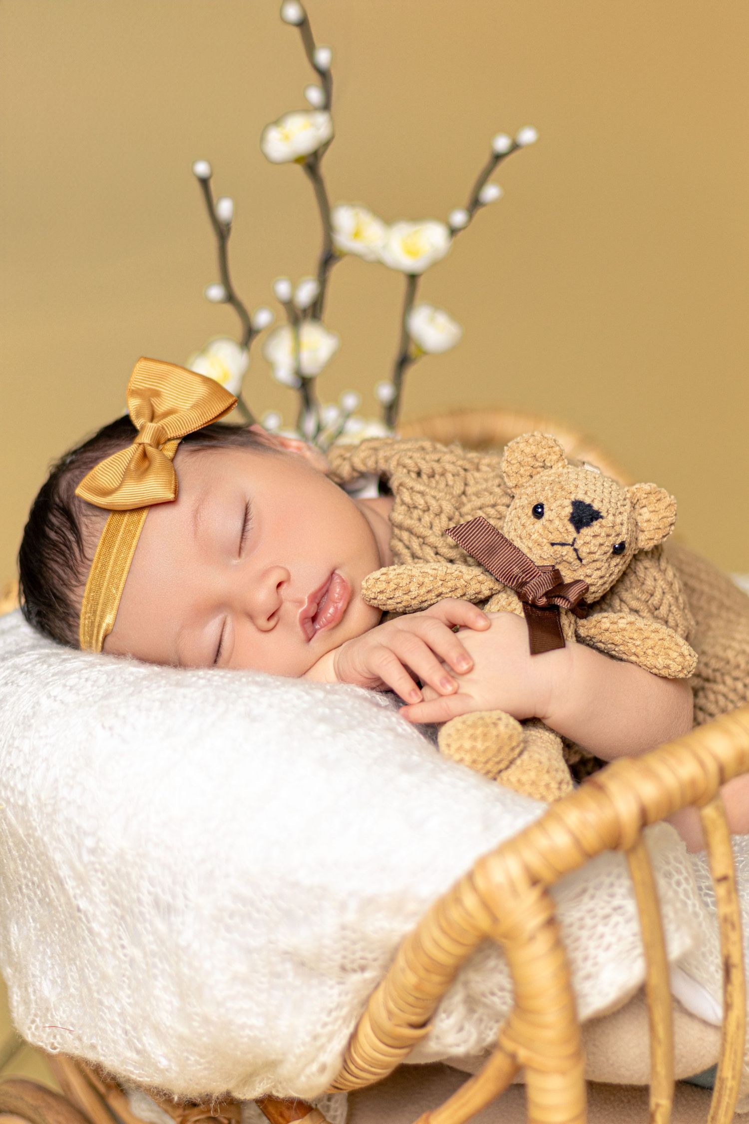 Sleeping newborn with knit teddy bear accessory and gold bow among white floral decorations.