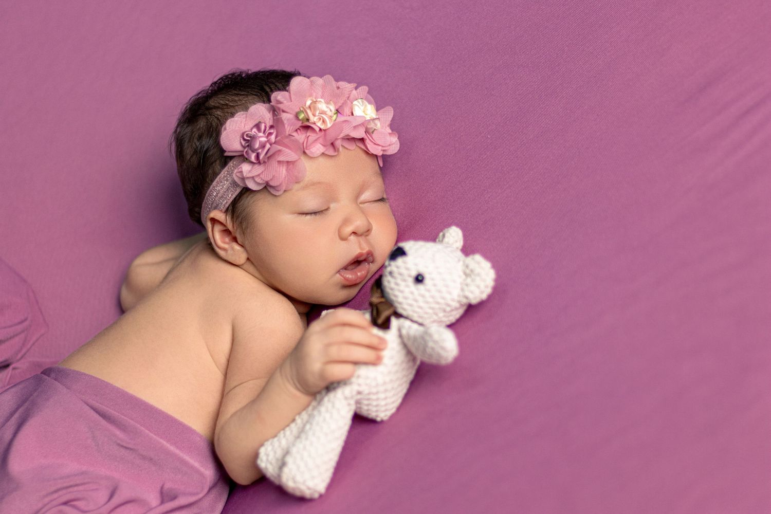 Sleeping newborn in artistic pose with white accessories against pink background.