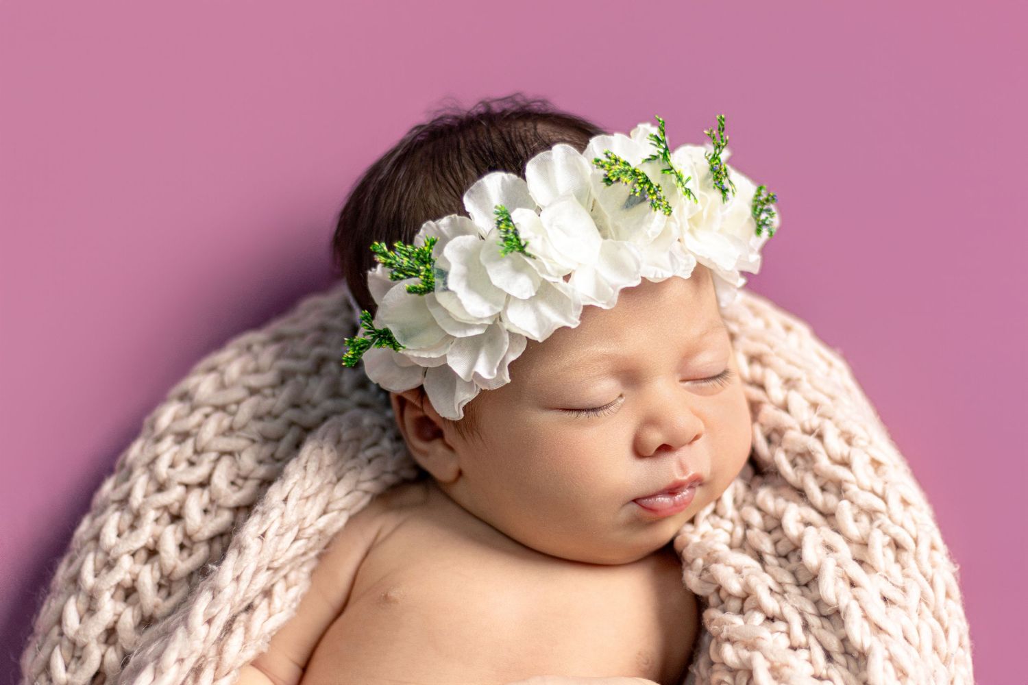Sleeping newborn wearing white floral crown against pink backdrop.