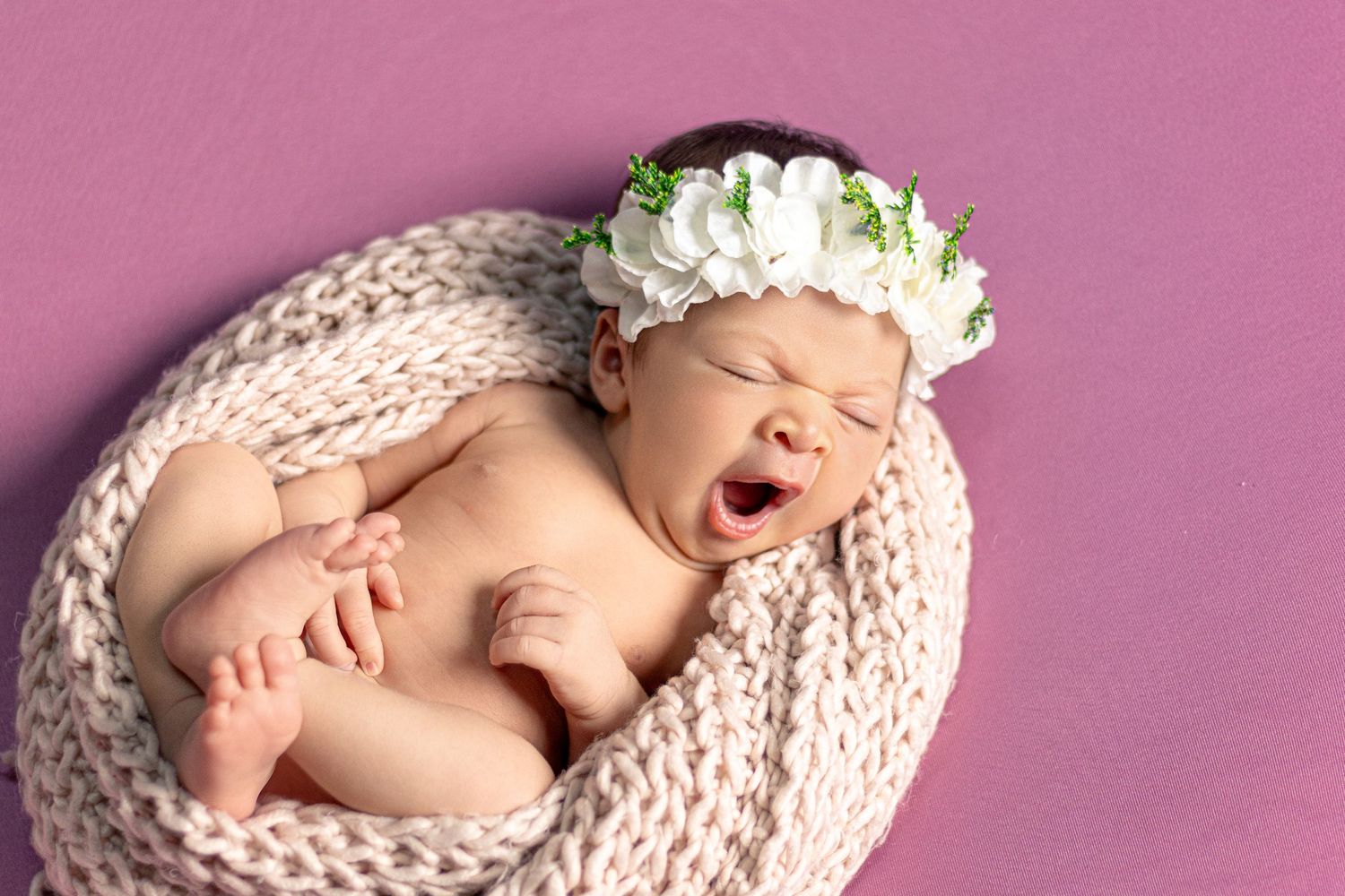 Peaceful newborn portrait with white flower crown and knit wrap on pink background.
