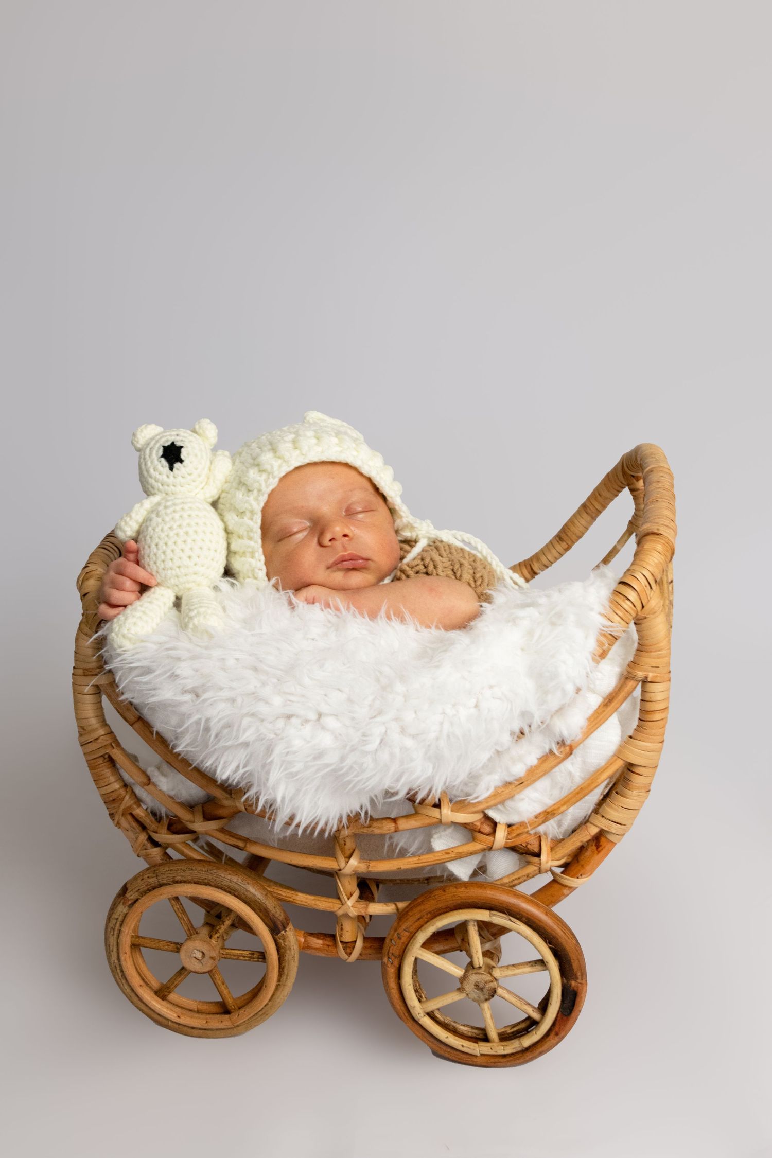 Sleeping newborn in white knit hat lying in vintage wicker carriage with plush toy.