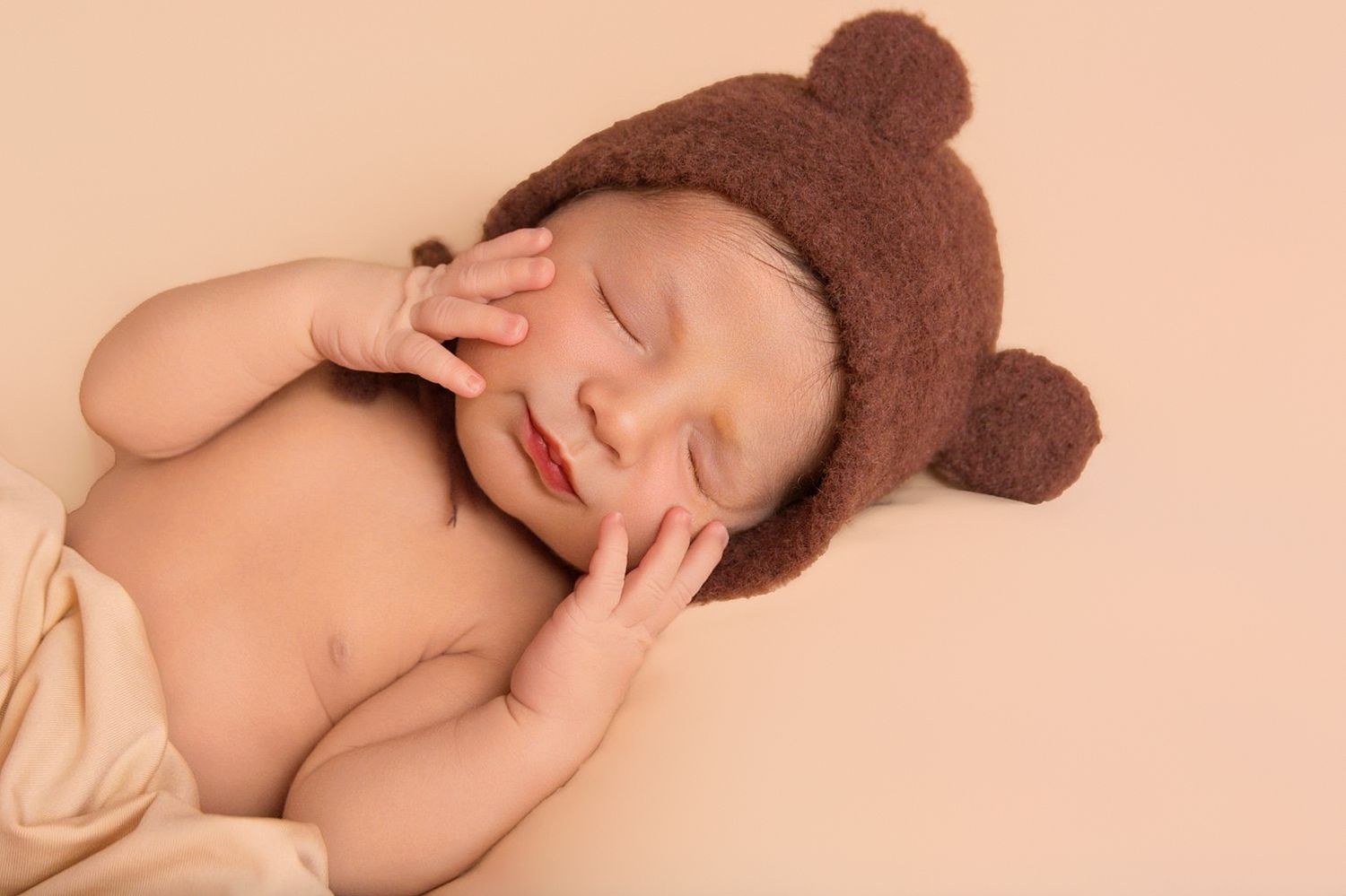 Sleeping newborn wearing brown teddy bear hat lies peacefully with hands near face.