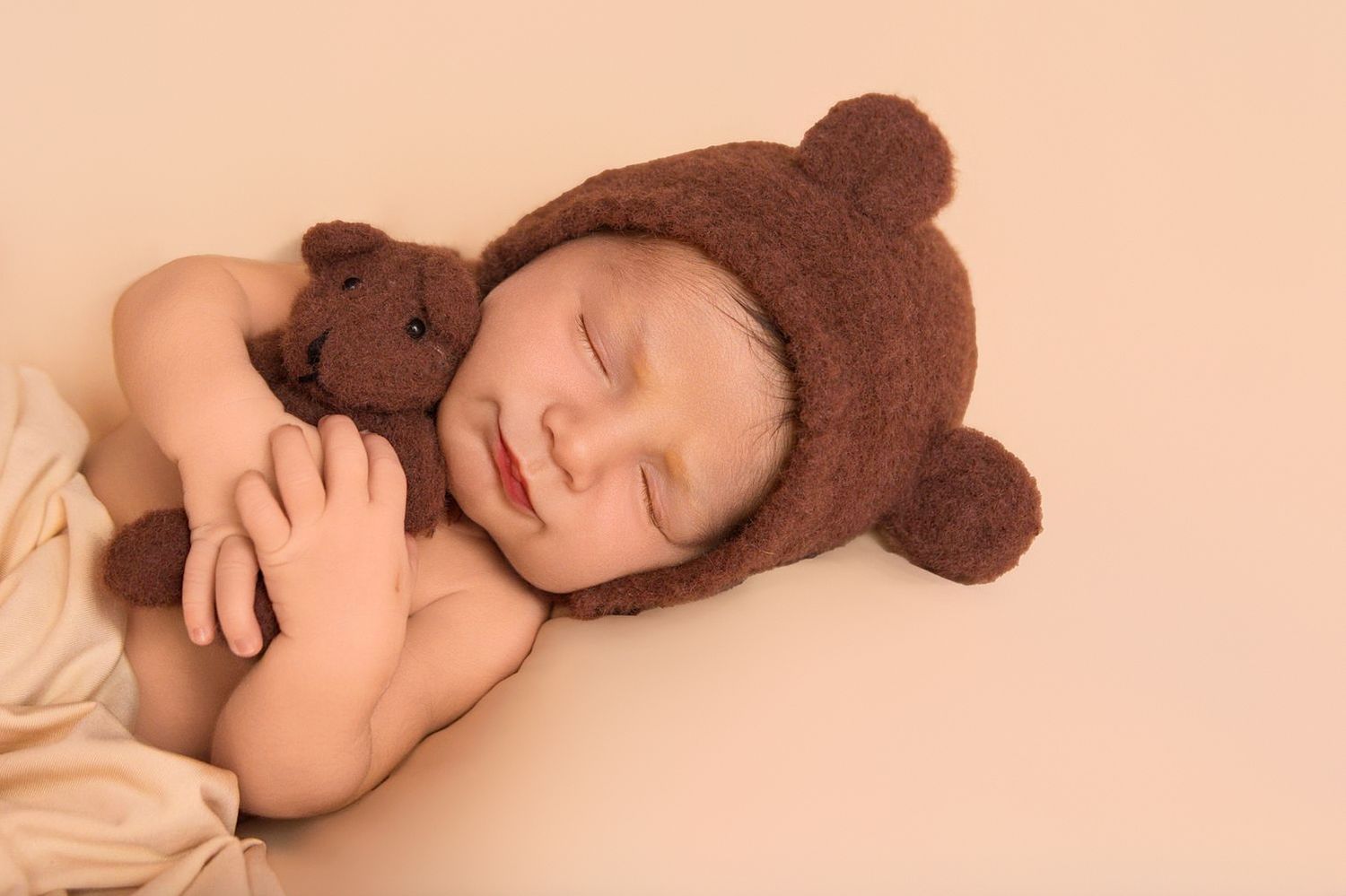 A sleeping baby lies on a cream-colored blanket wearing a brown teddy bear hat.