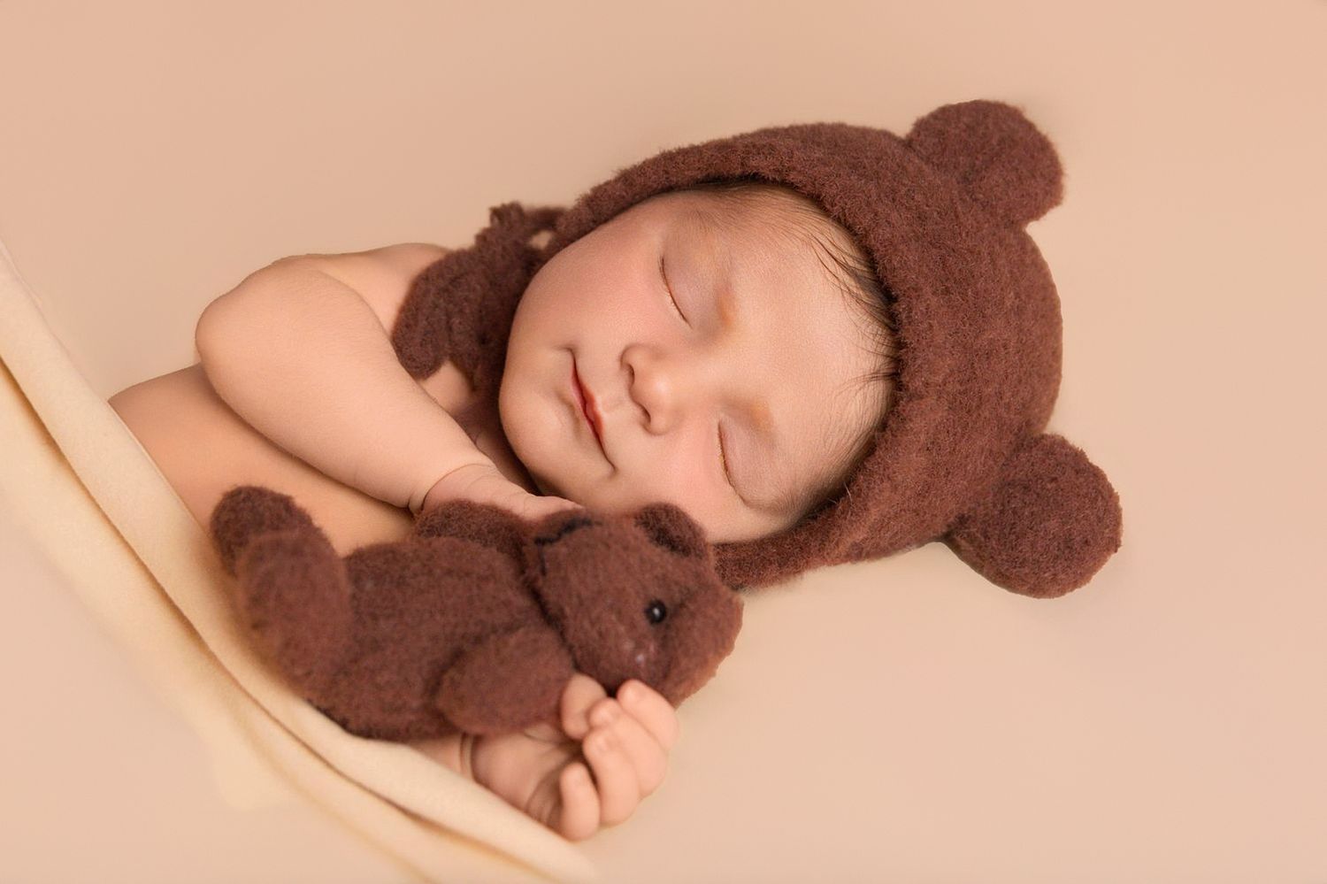A sleeping baby wearing a brown bear hat lies peacefully with a matching teddy bear on a cream-colored backdrop.