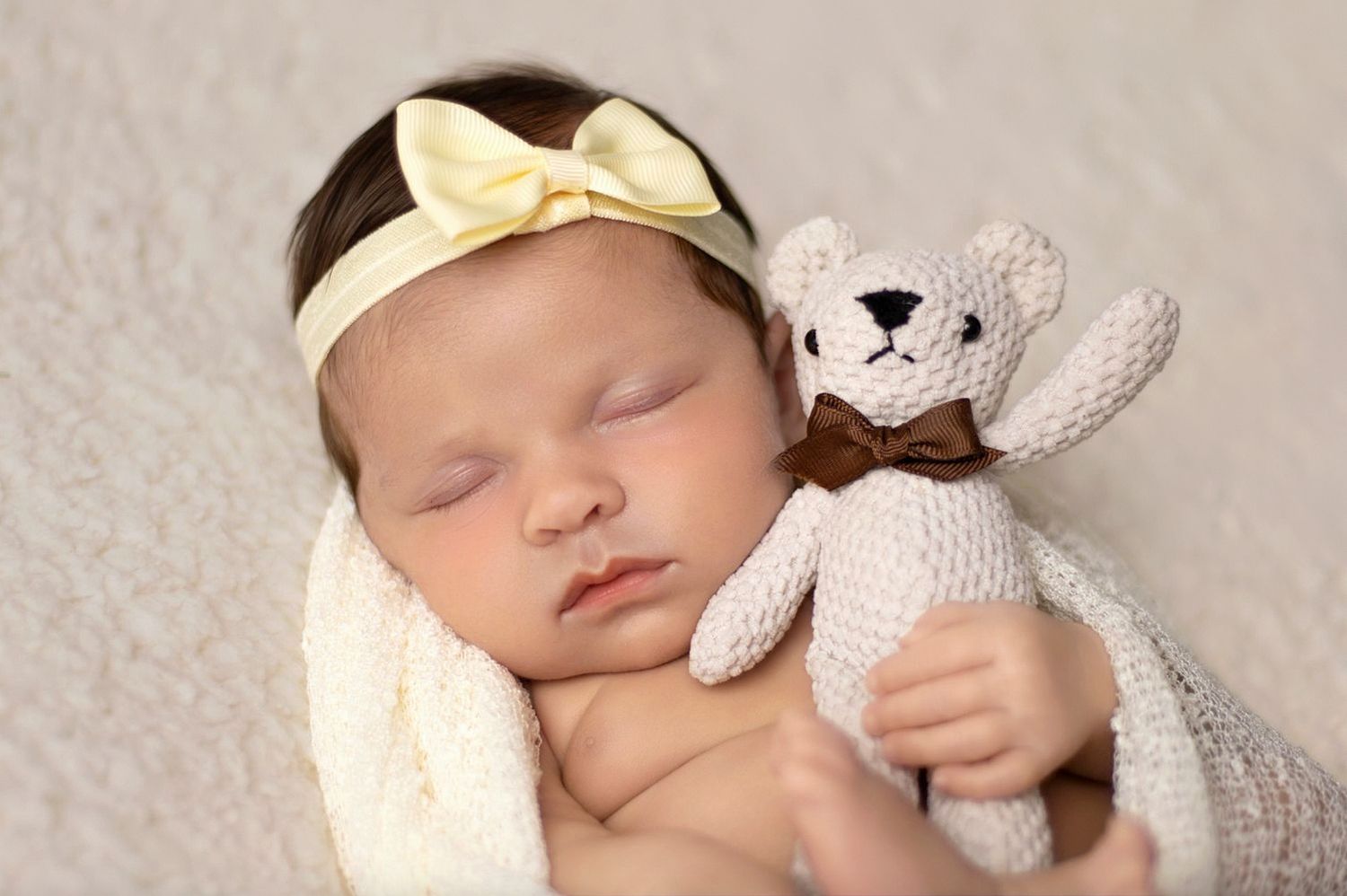 A sleeping baby wearing a white bow headband cuddles with a small knitted bunny toy on cream-colored bedding.