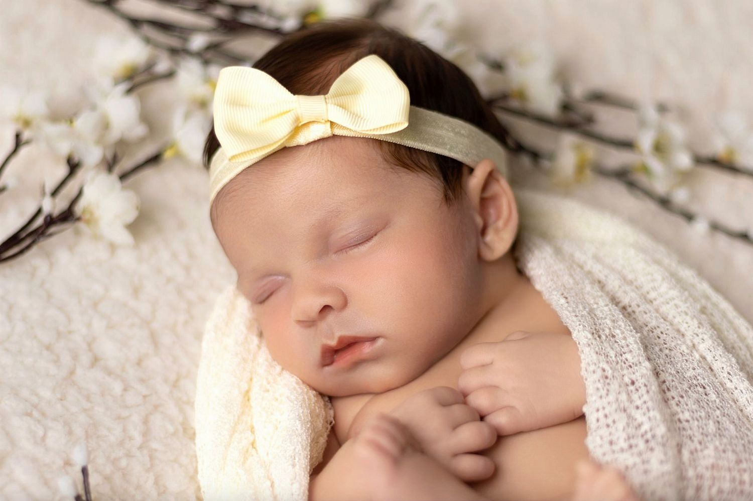 Peaceful sleeping newborn wearing cream bow headband wrapped in soft white knit blanket beside willow branches.