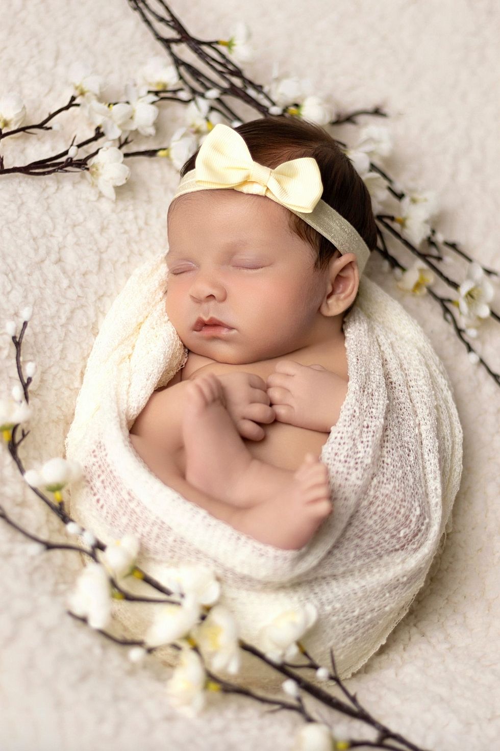 Sleeping newborn wrapped in white knit blanket surrounded by delicate white cherry blossoms and wearing cream bow headband.