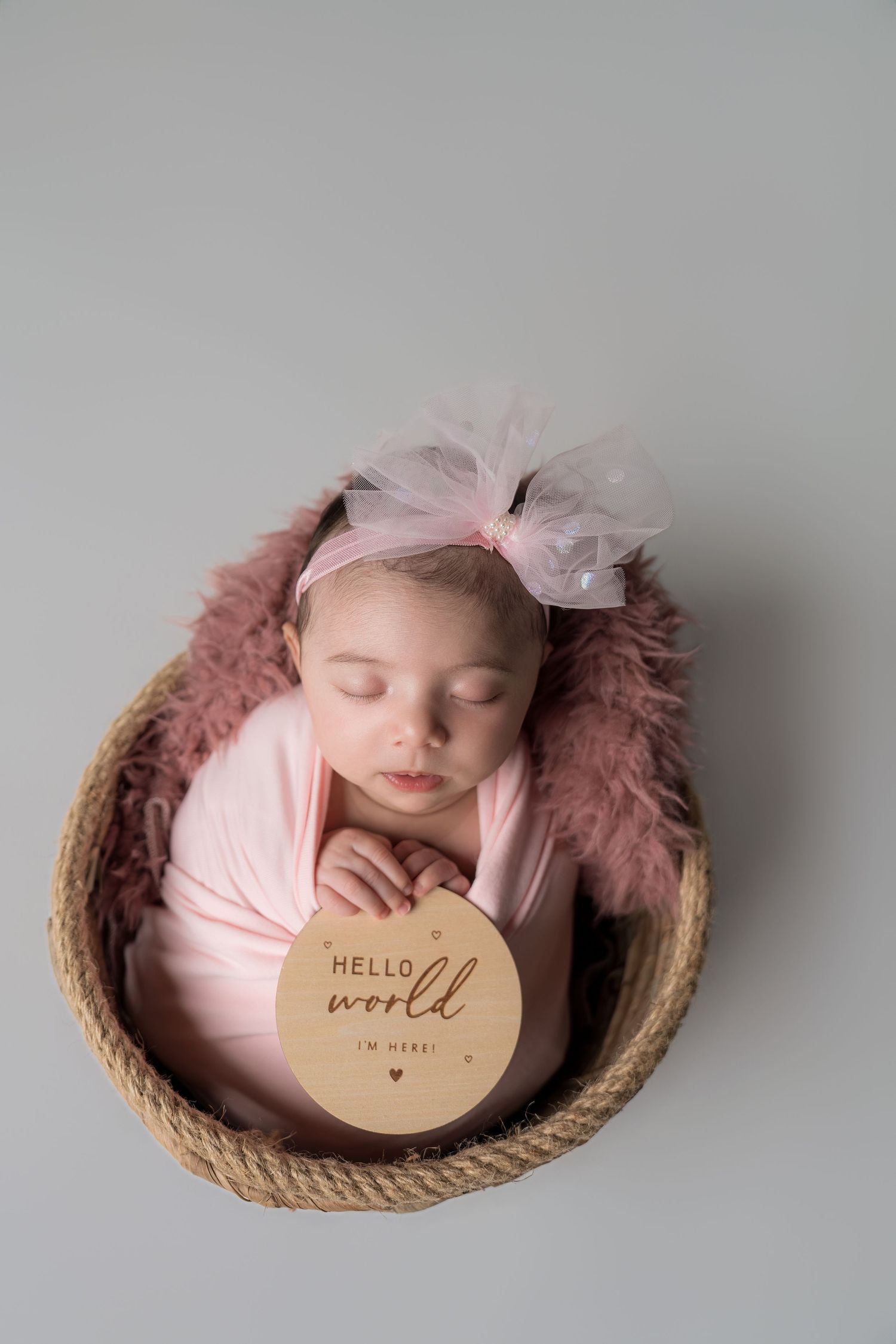 Newborn baby sleeping peacefully in round basket with pink blanket and bow, next to 'Hello World' wooden sign.