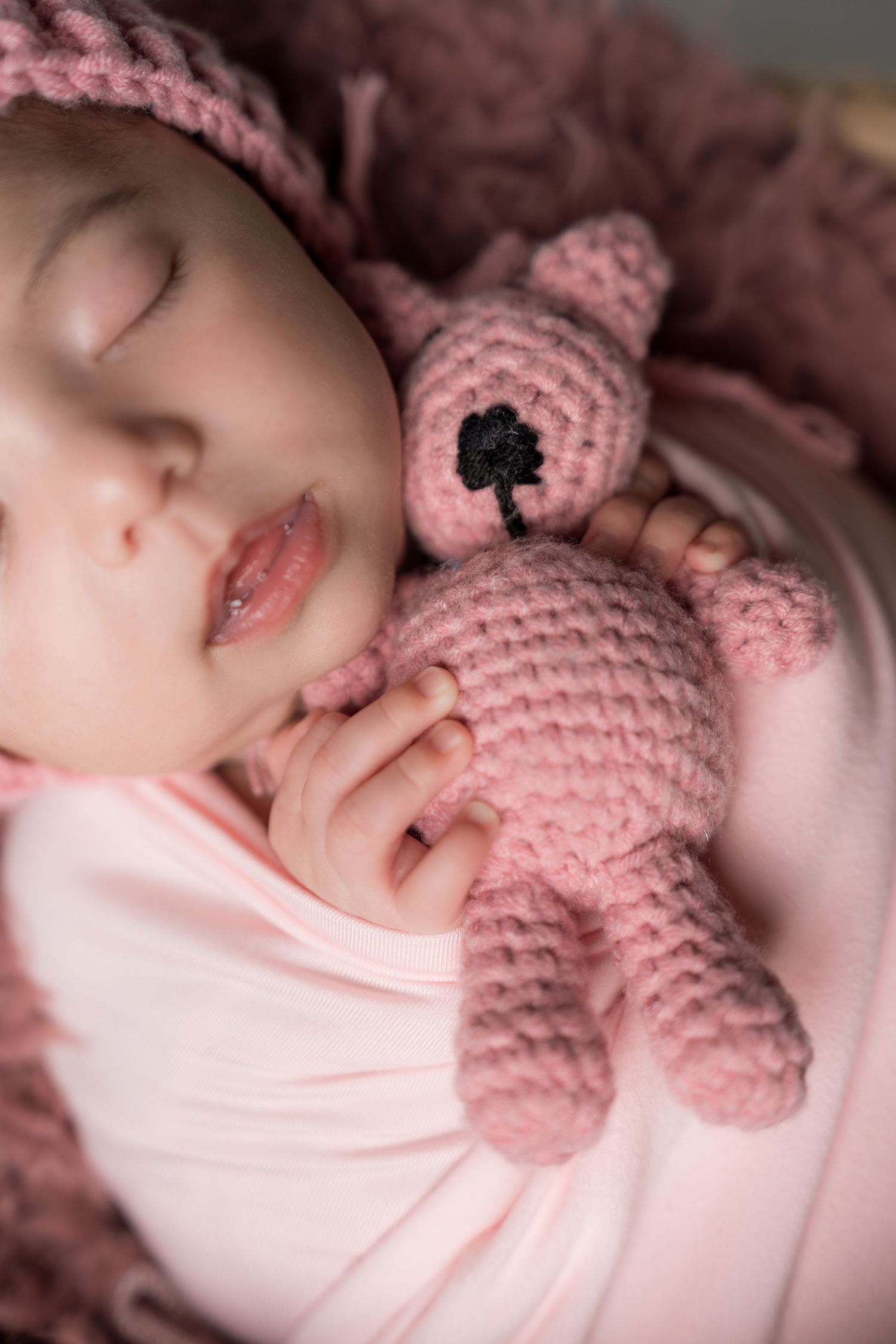 Sleeping baby cuddling with pink knitted teddy bear while wearing matching pink hat.