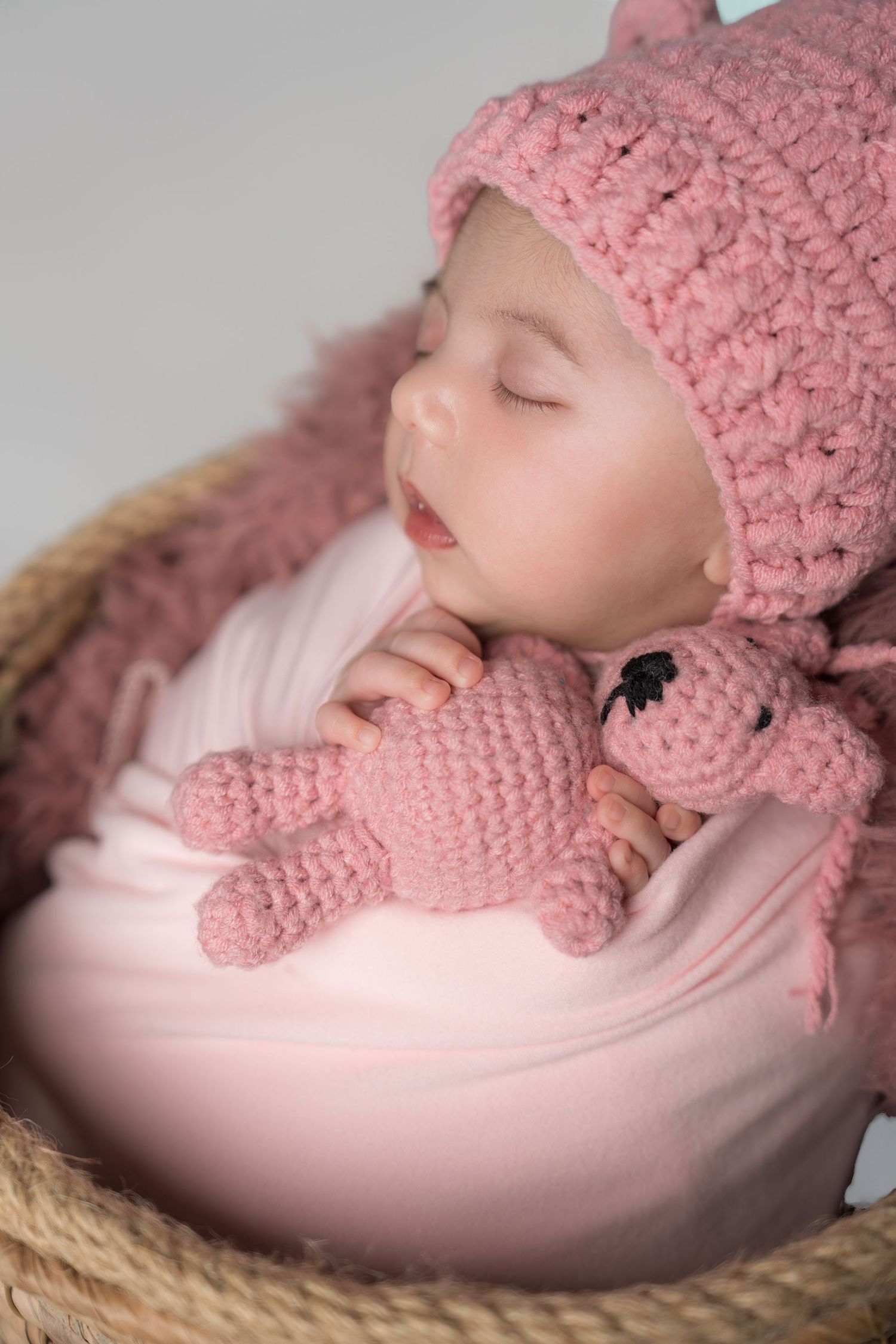 A sleeping baby wearing a pink knitted hat rests peacefully in a cozy basket with soft lighting.
