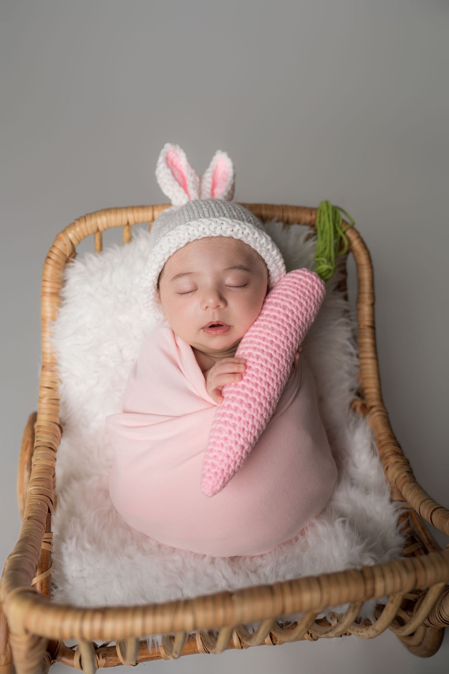 A sleeping newborn swaddled in pink with bunny ears rests peacefully in a wicker basket with white fluff bedding.