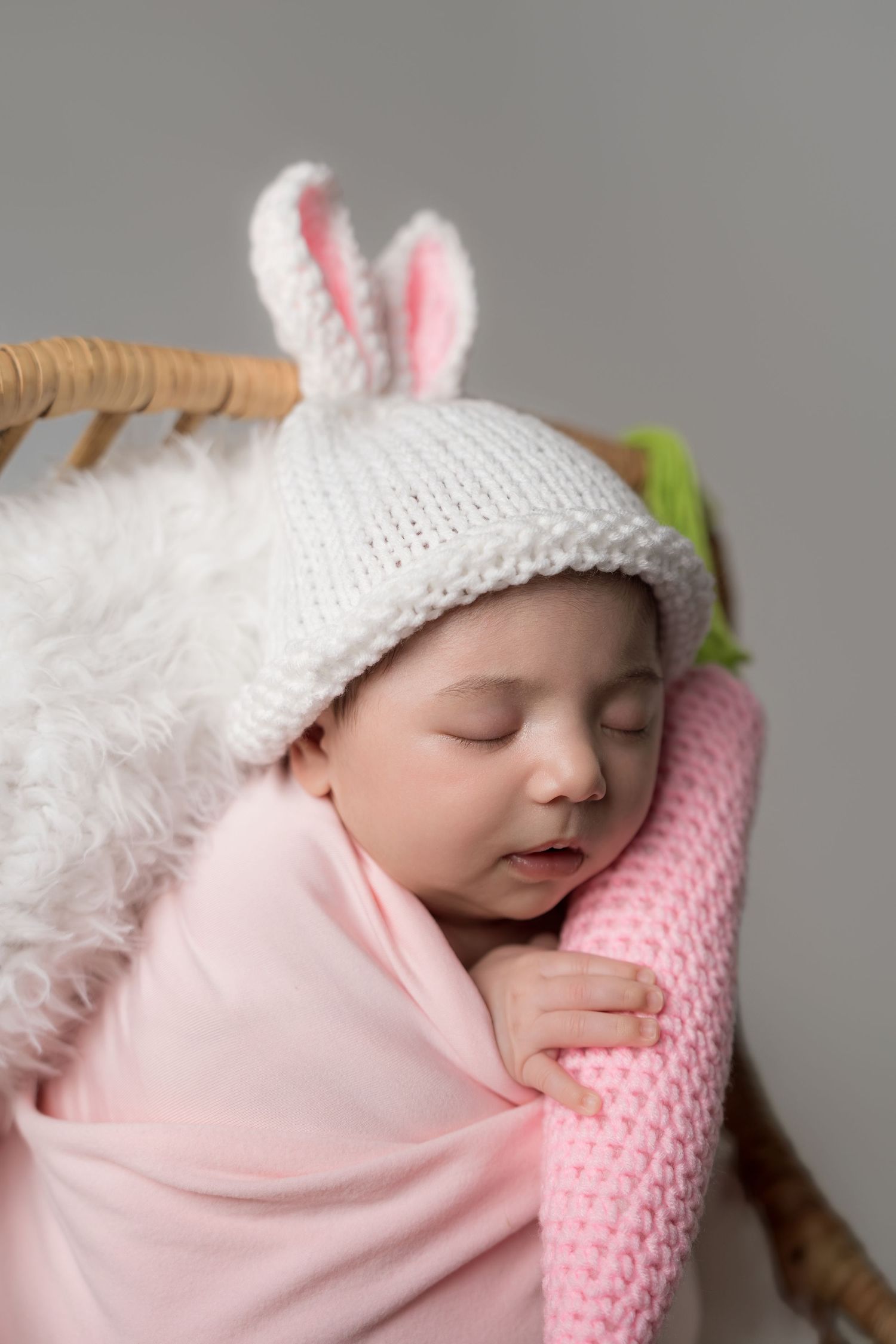 Sleeping baby wrapped in pink blanket wearing white knit hat with bunny ears rests on pink pillow.