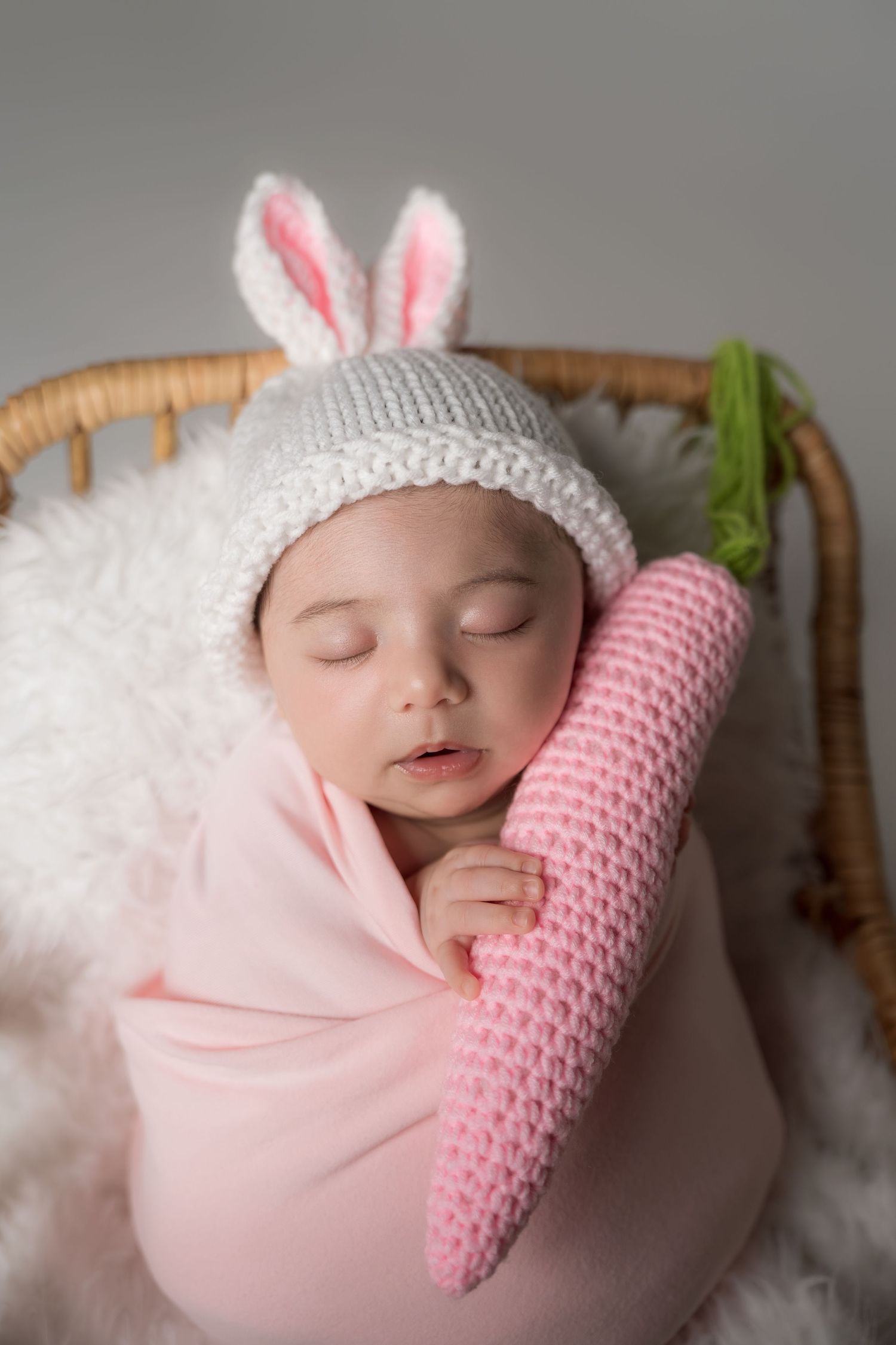 A sleeping baby wearing bunny ears rests peacefully on soft white bedding with a pink knitted pillow.