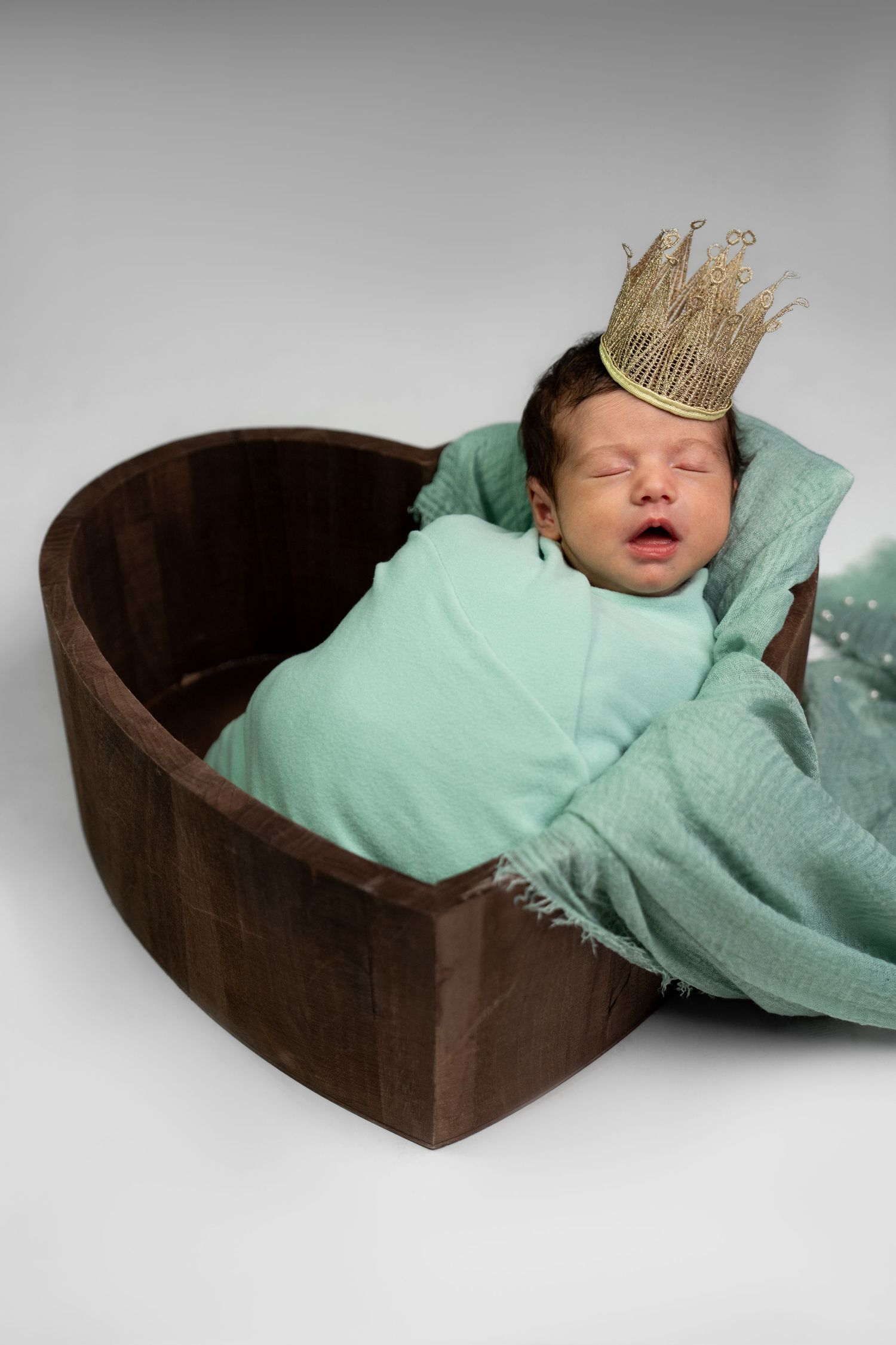 Newborn baby wearing a small crown sleeps in a brown basket with mint green blanket.