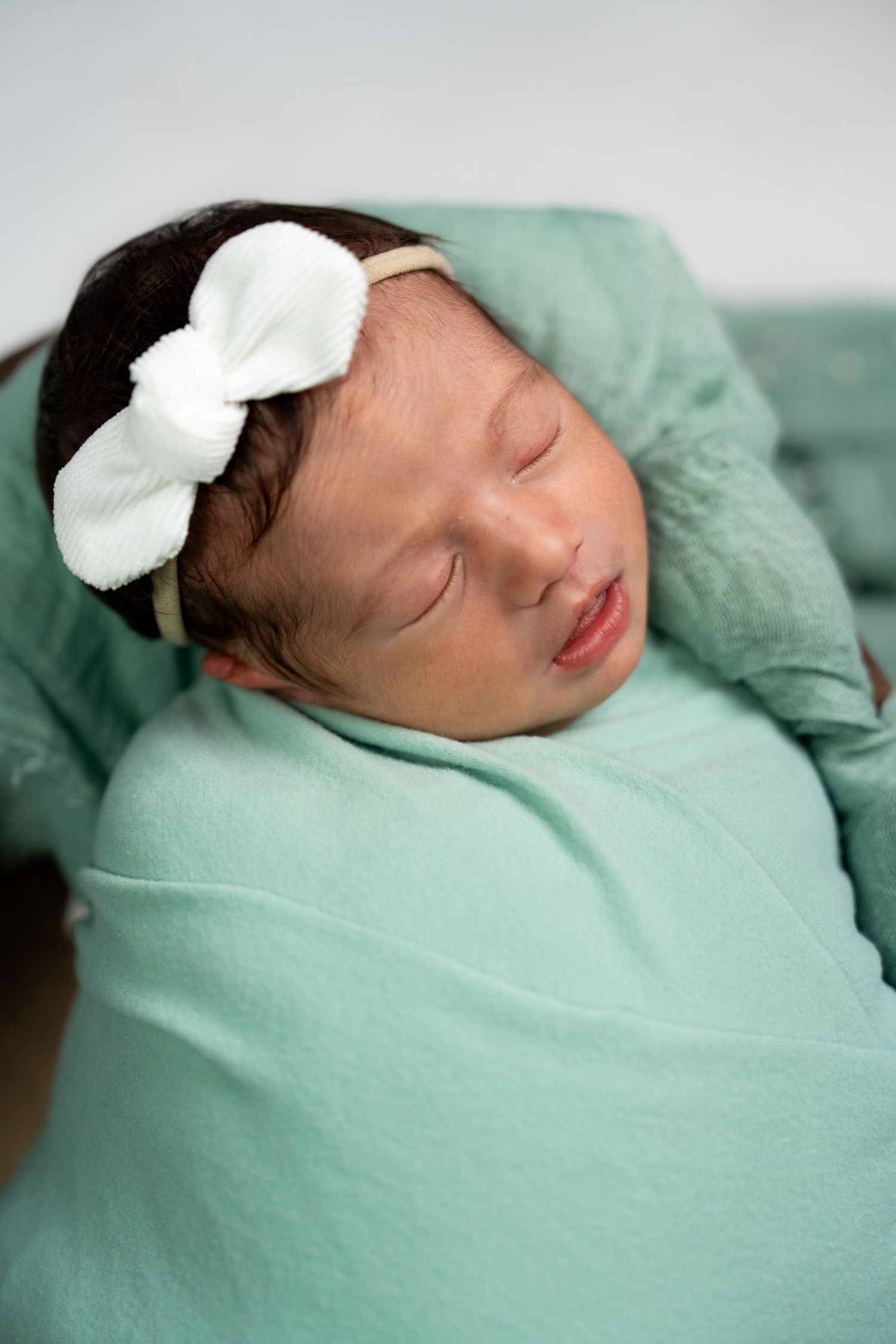 Sleeping newborn with white bow accessory wrapped in soft mint green fabric.