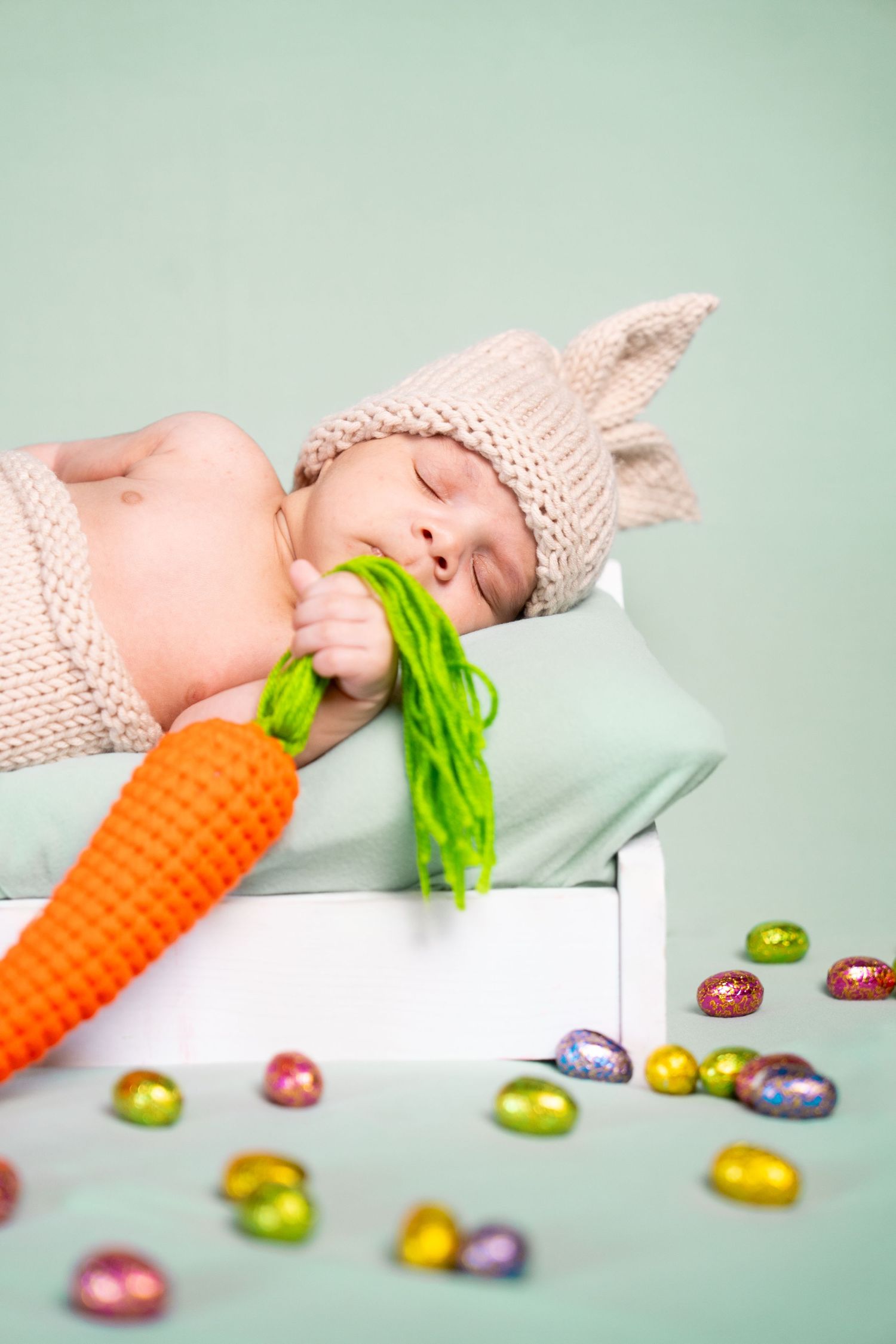 Sleeping newborn in bunny outfit with orange carrot prop and scattered Easter eggs.