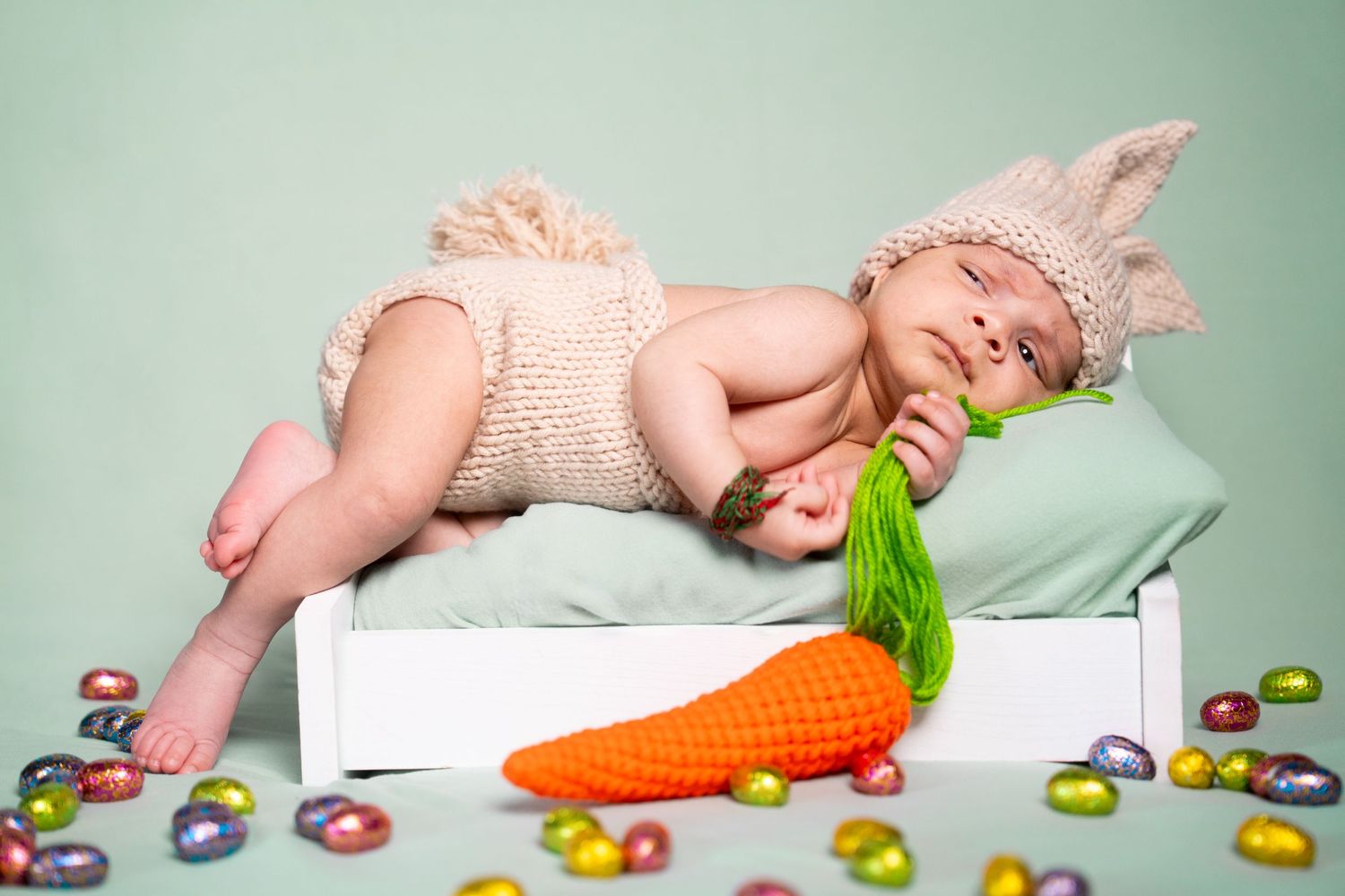 Baby in bunny costume with carrot prop surrounded by colorful Easter eggs on mint background.