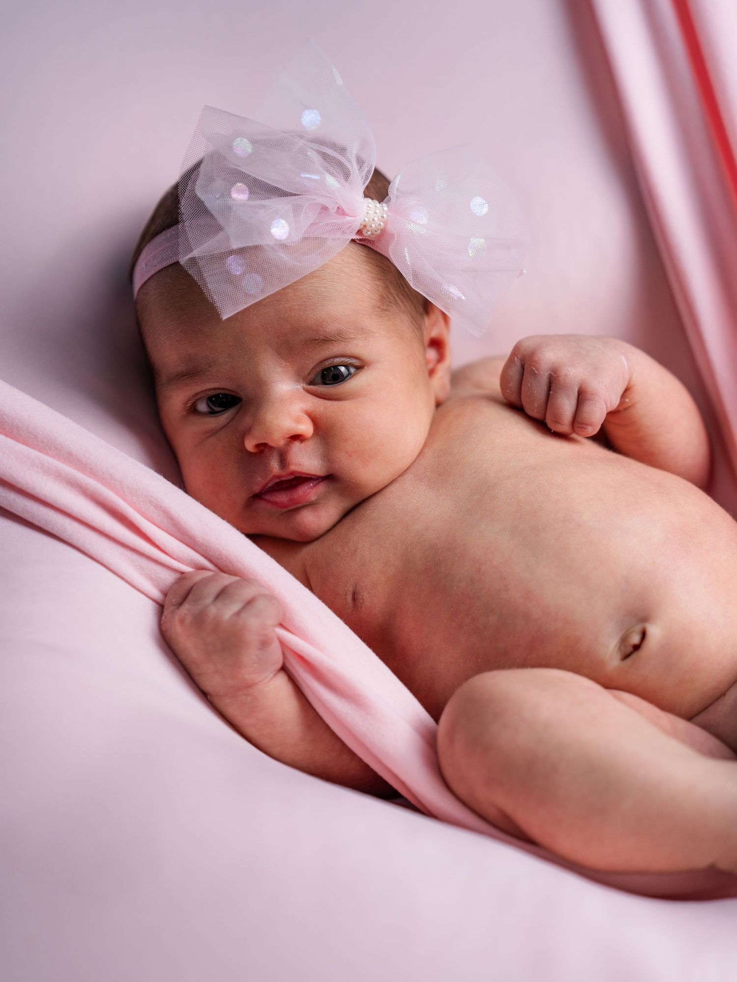 Newborn baby wearing white bow lying on pink bedding.