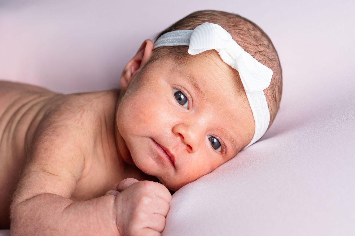Side view of newborn baby with white bow headband on purple background.