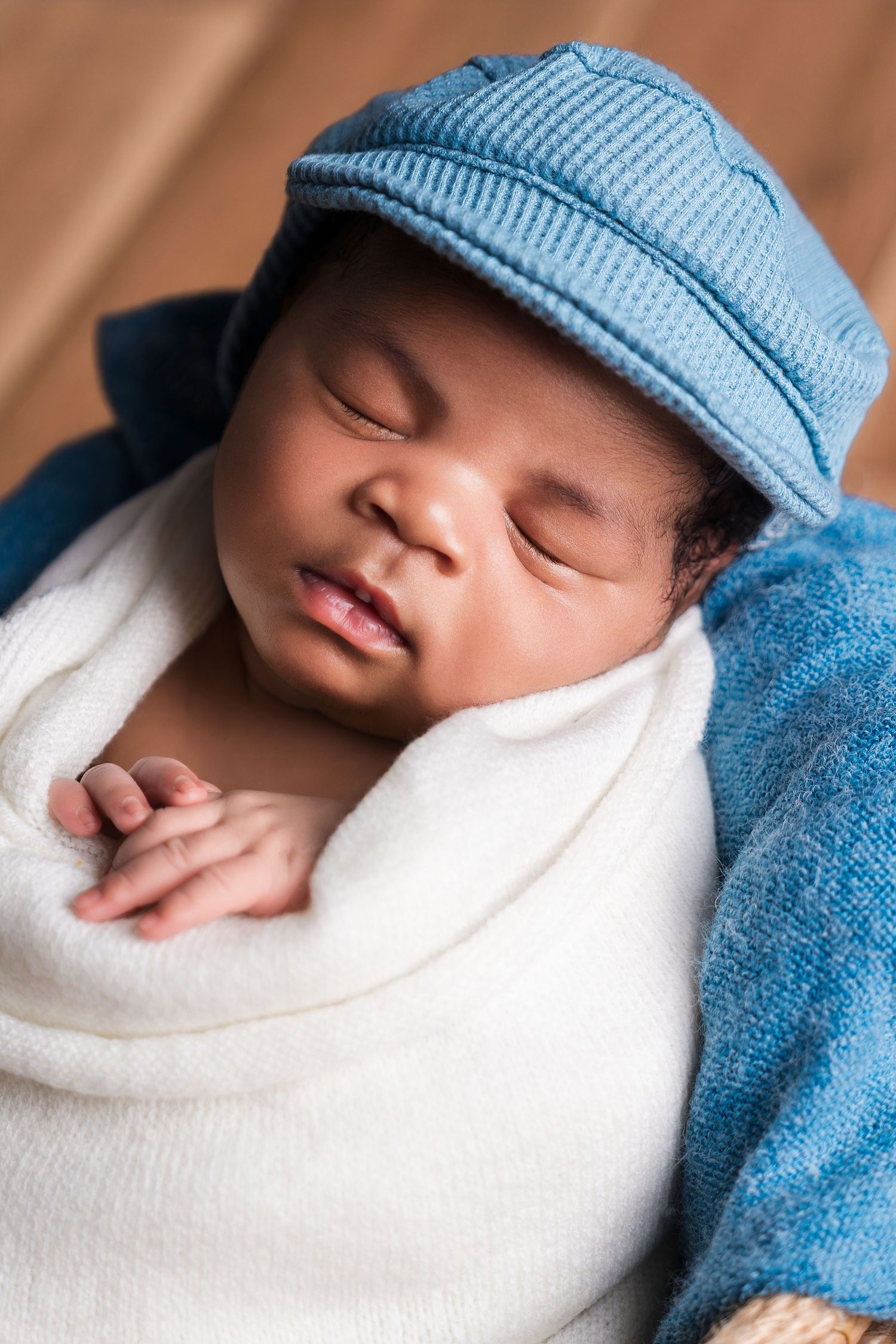 Sleeping newborn baby wearing a blue newsboy cap, swaddled in a white blanket with a blue wrap.