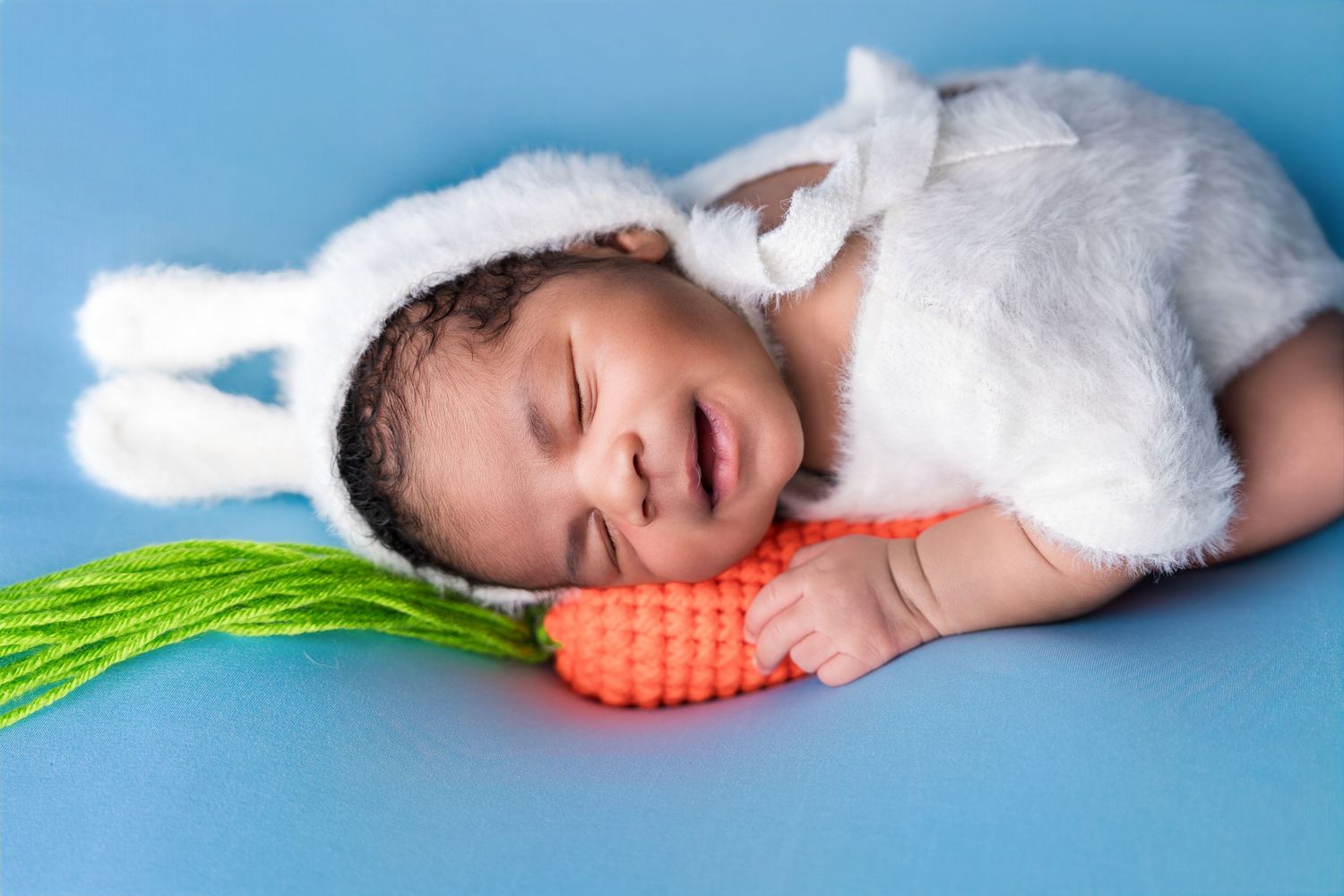 Newborn baby sleeping in white bunny costume with ears, holding a toy carrot on blue background.