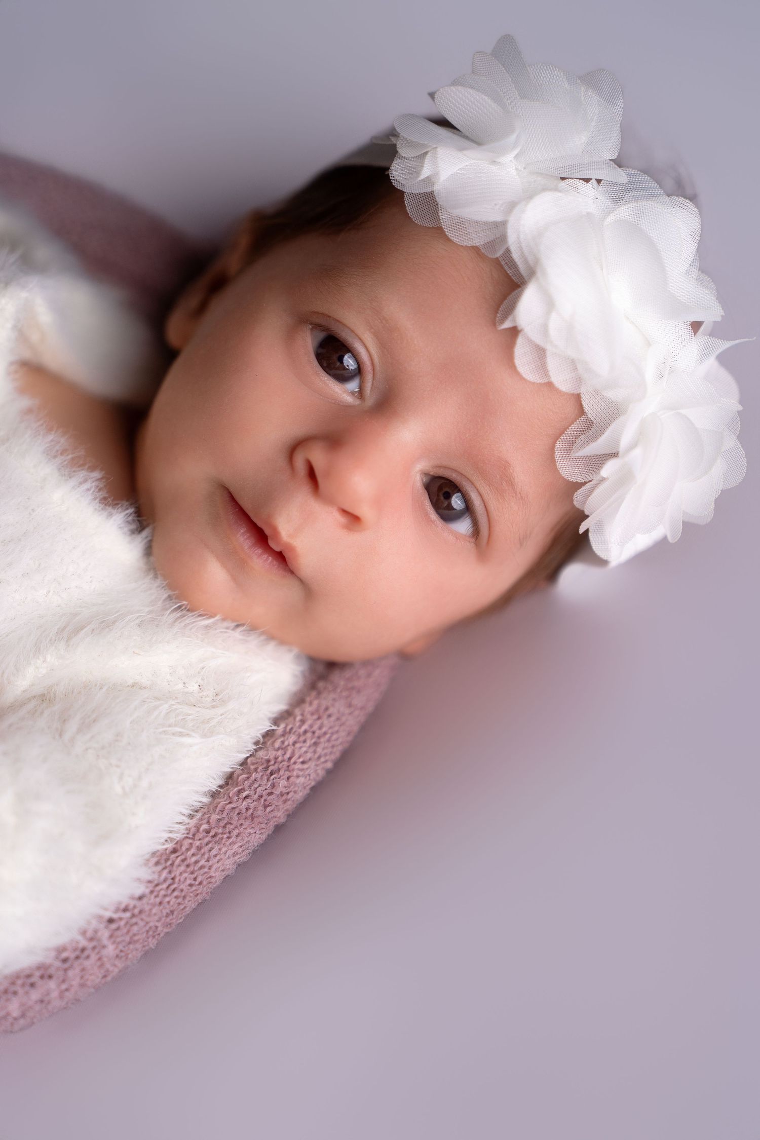 Baby wearing white floral headband lying on soft fabric in studio setting.
