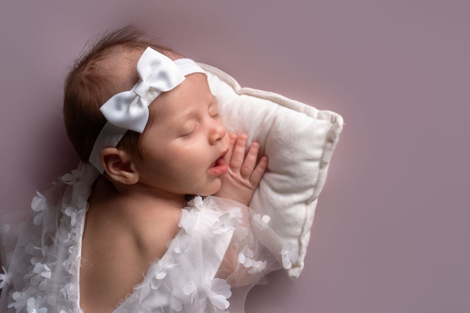 Sleeping baby in white lace outfit on pink studio backdrop.