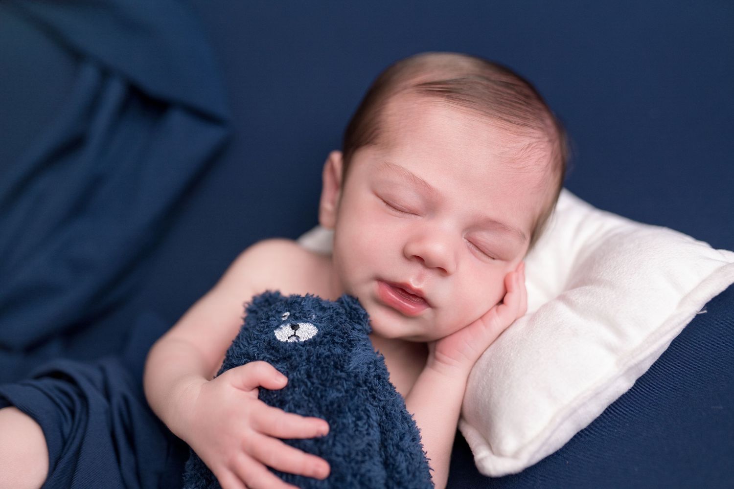 Peaceful newborn baby sleeping on a white pillow, cuddling a navy blue teddy bear, wrapped in a soft blue blanket.
